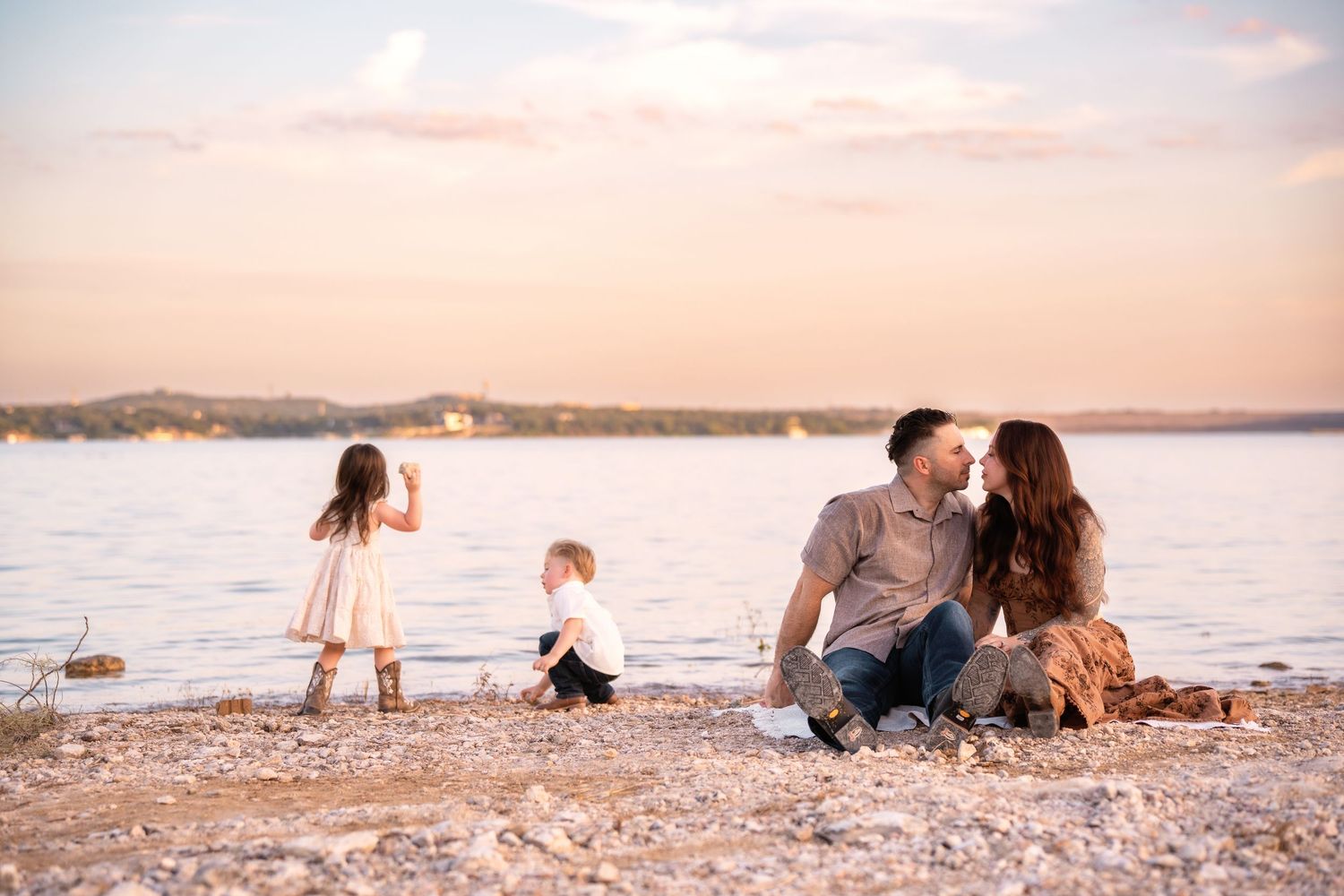 Family relaxing on rocky beach shoreline during sunset with water and mountains in background.