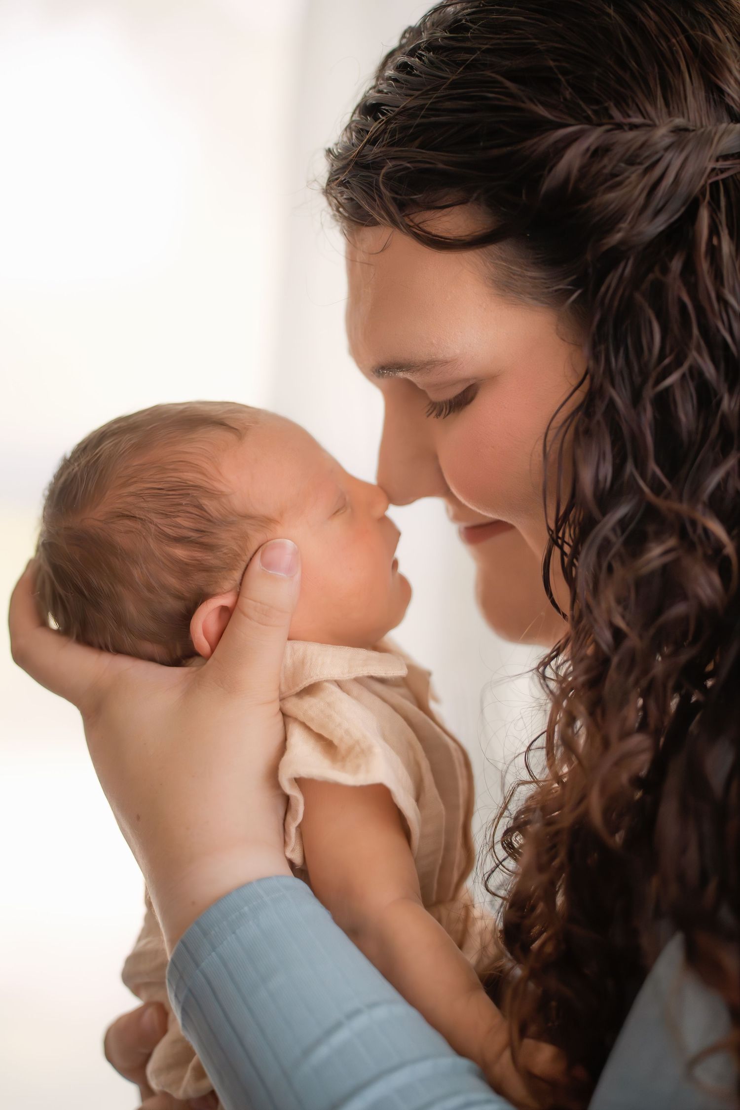 A tender moment as a curly-haired mother gently touches noses with her newborn baby cradled in her hands.