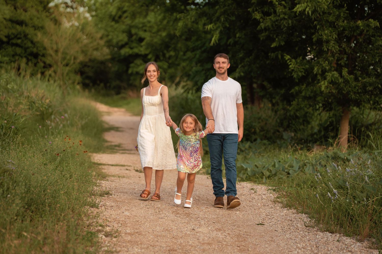 A young family of three walks hand-in-hand on a sunny gravel path surrounded by lush green trees.