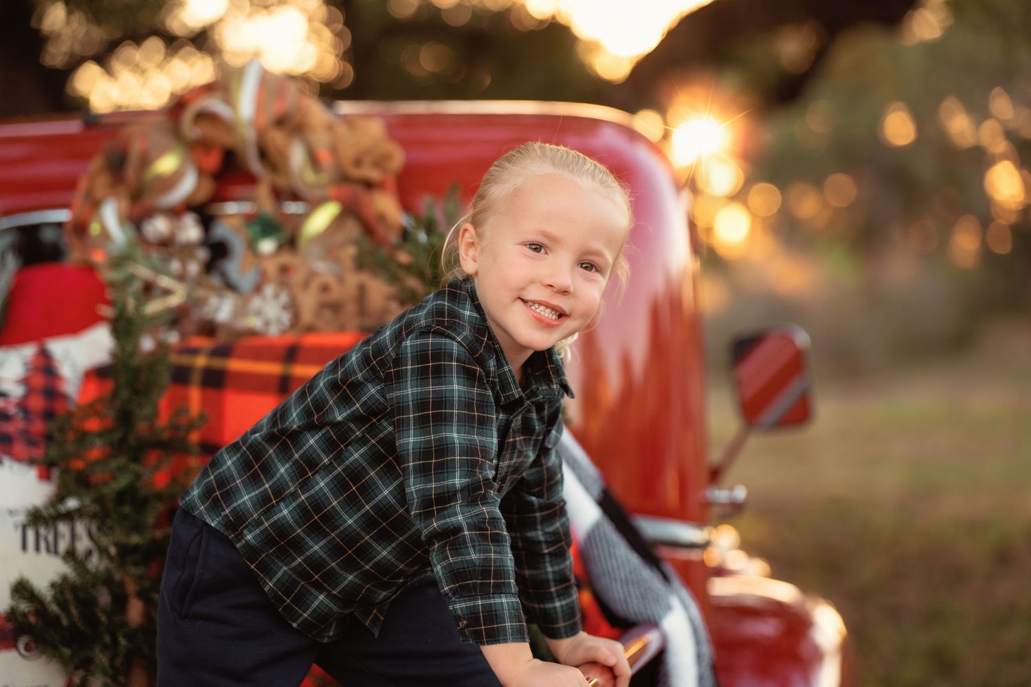 A smiling young boy in a plaid shirt poses beside a decorated red vintage truck with Christmas lights outdoors.