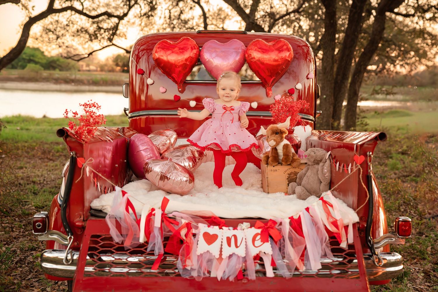 Baby girl in a pink dress sits in a vintage red truck bed decorated with heart balloons, teddy bears, and Valentine's Day banners outdoors.