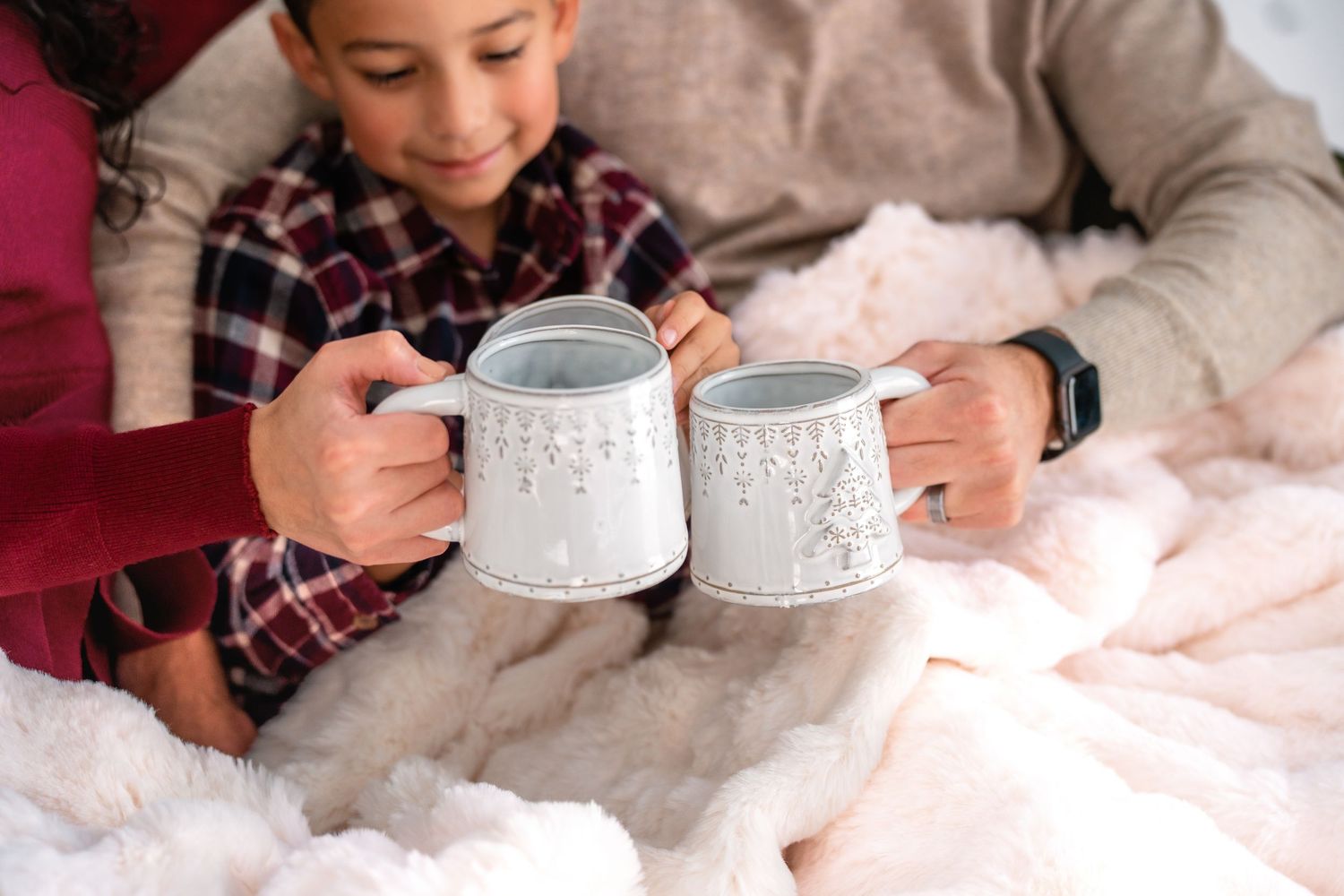 Coffee mugs held close together in a festive holiday setting with white decor and soft lighting.