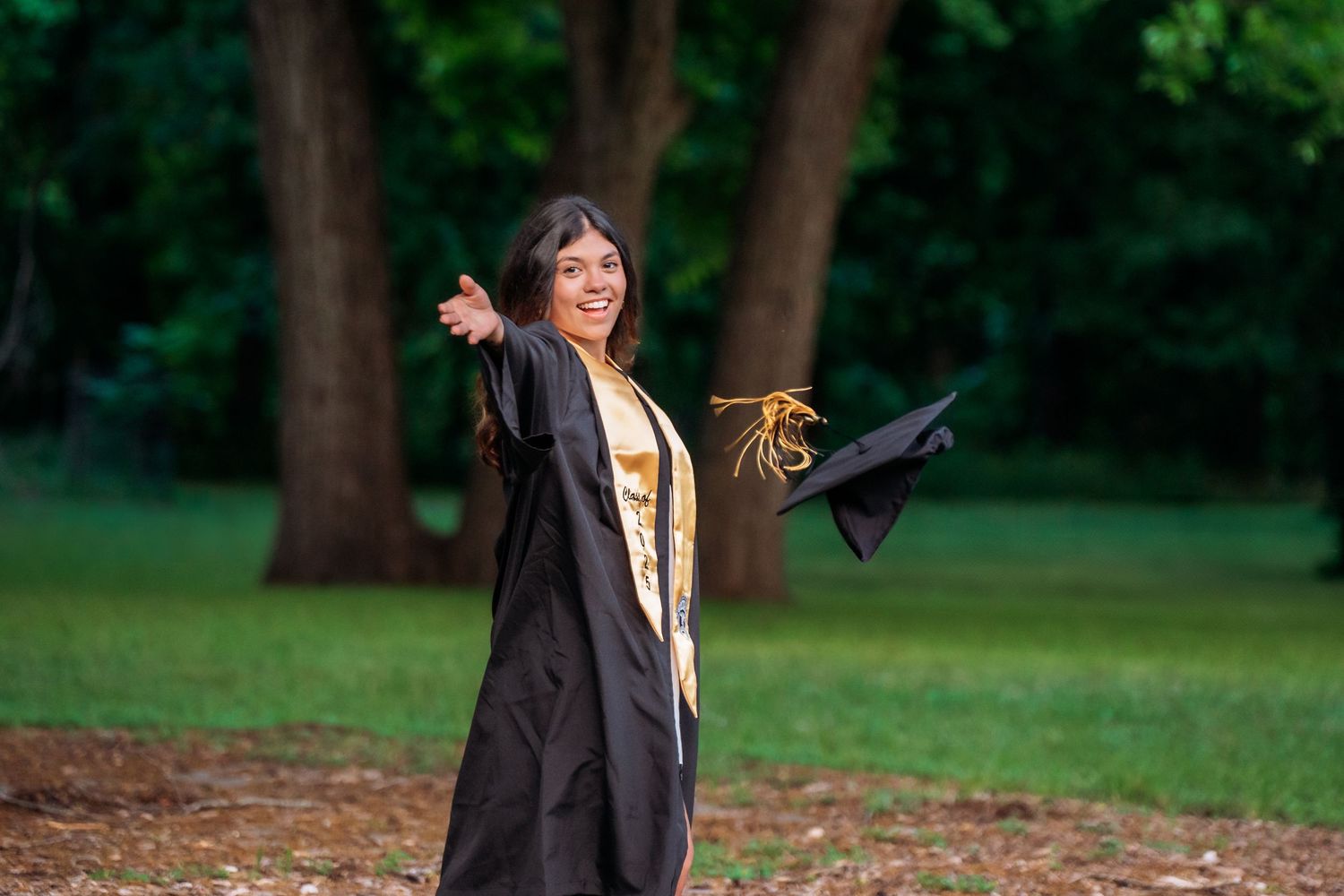 Young woman in black and gold graduation gown tossing her cap in a lush green park, celebrating her achievement.