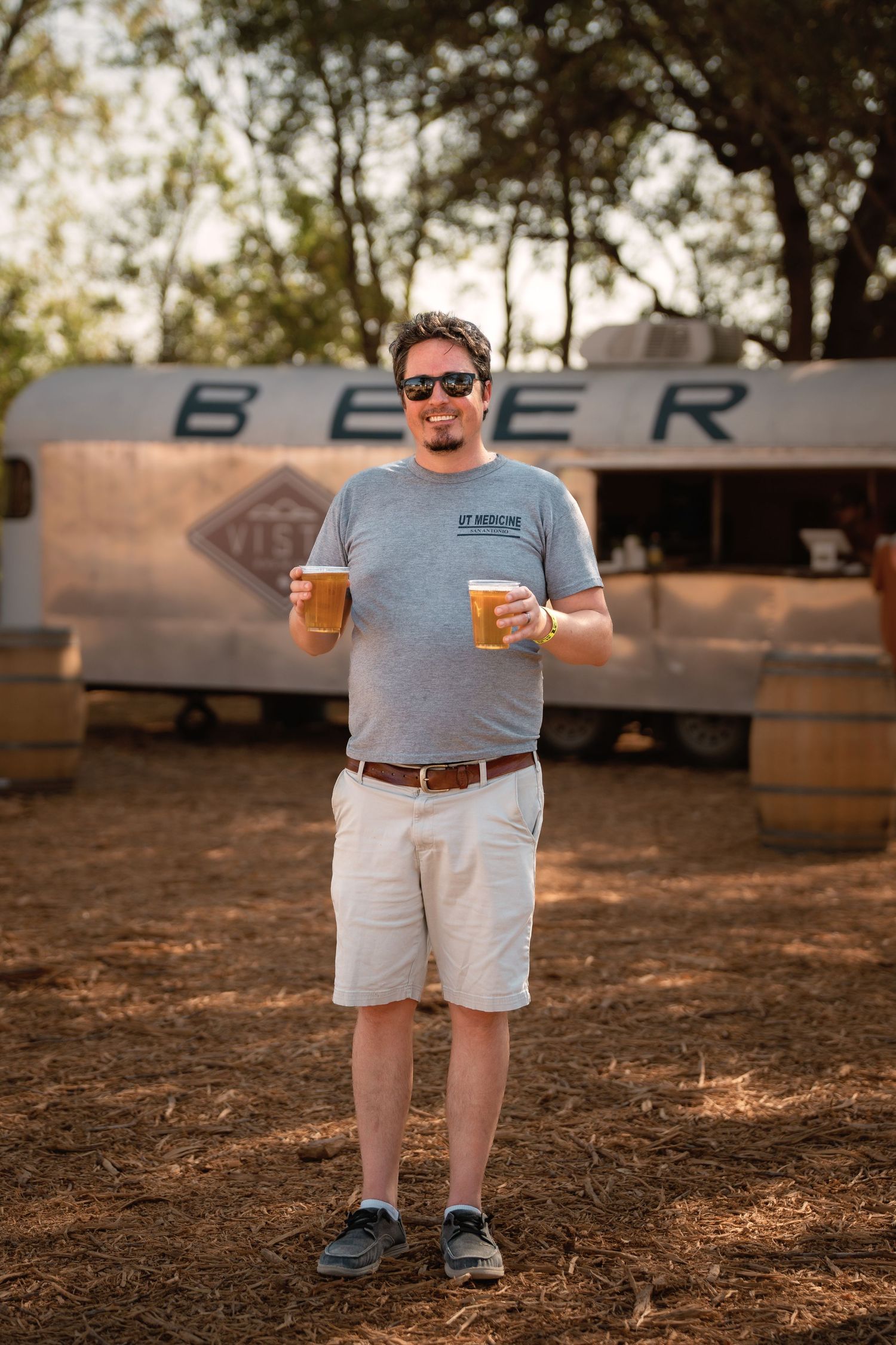 Man holding two beers at outdoor festival with vintage trailer beer stand in background.