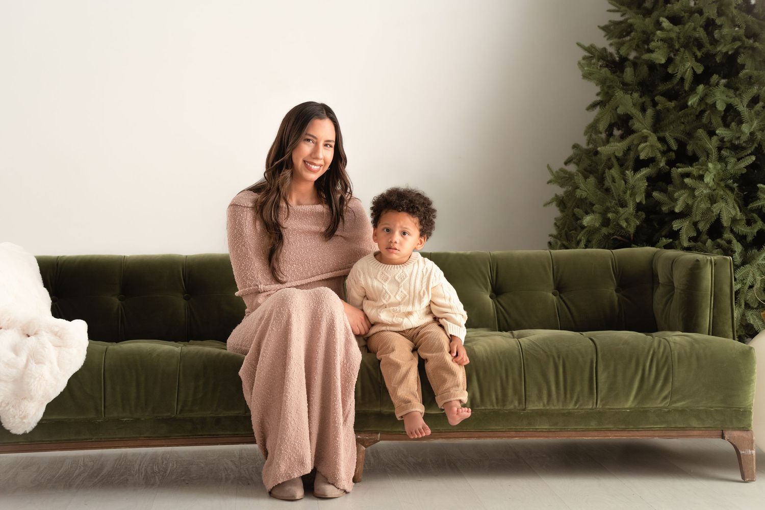 Woman and toddler sitting on green velvet sofa near Christmas tree, wearing neutral tones for holiday photo.