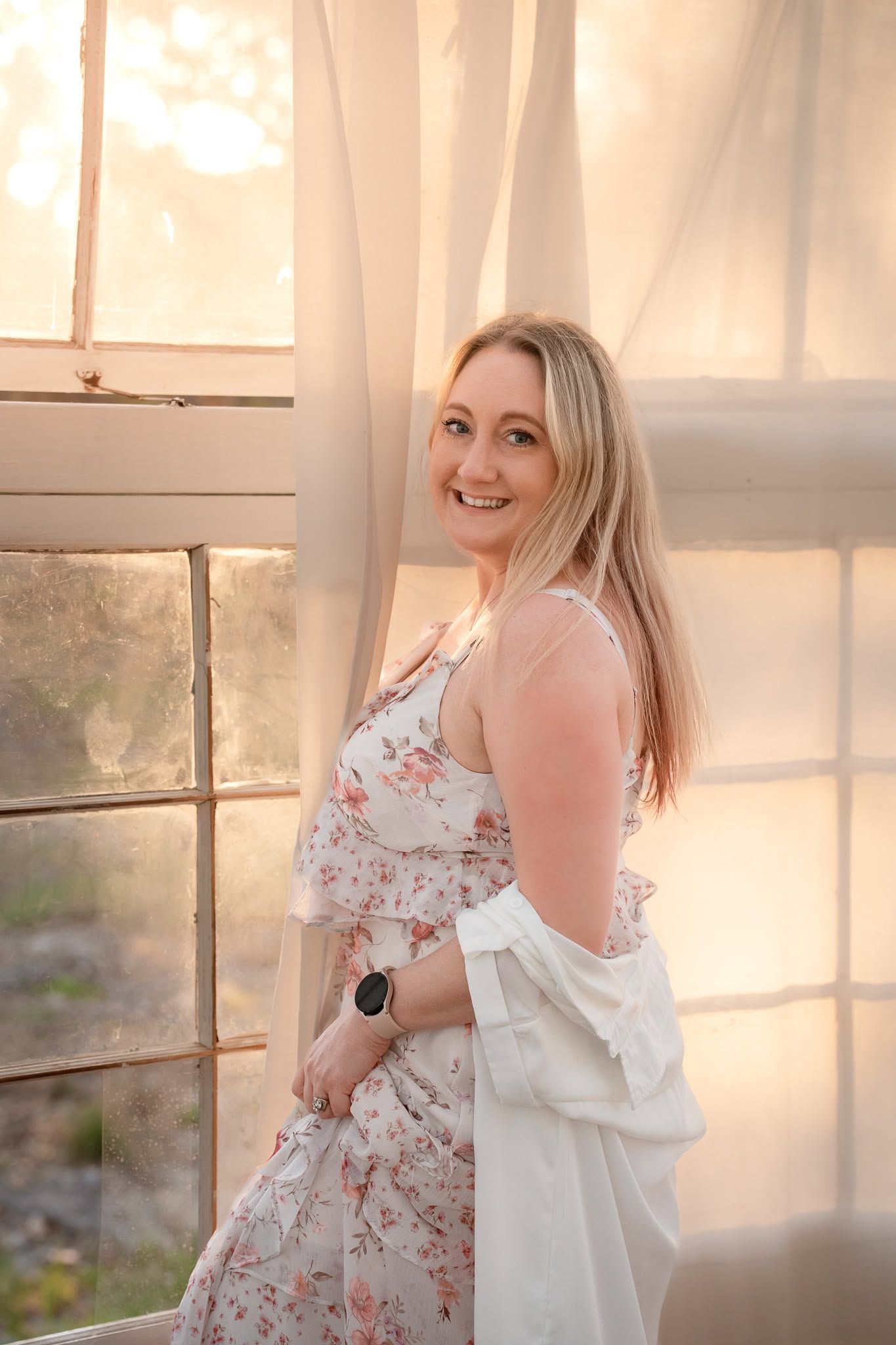 Smiling blonde pregnant woman in floral dress and white jacket poses by sunlit window.