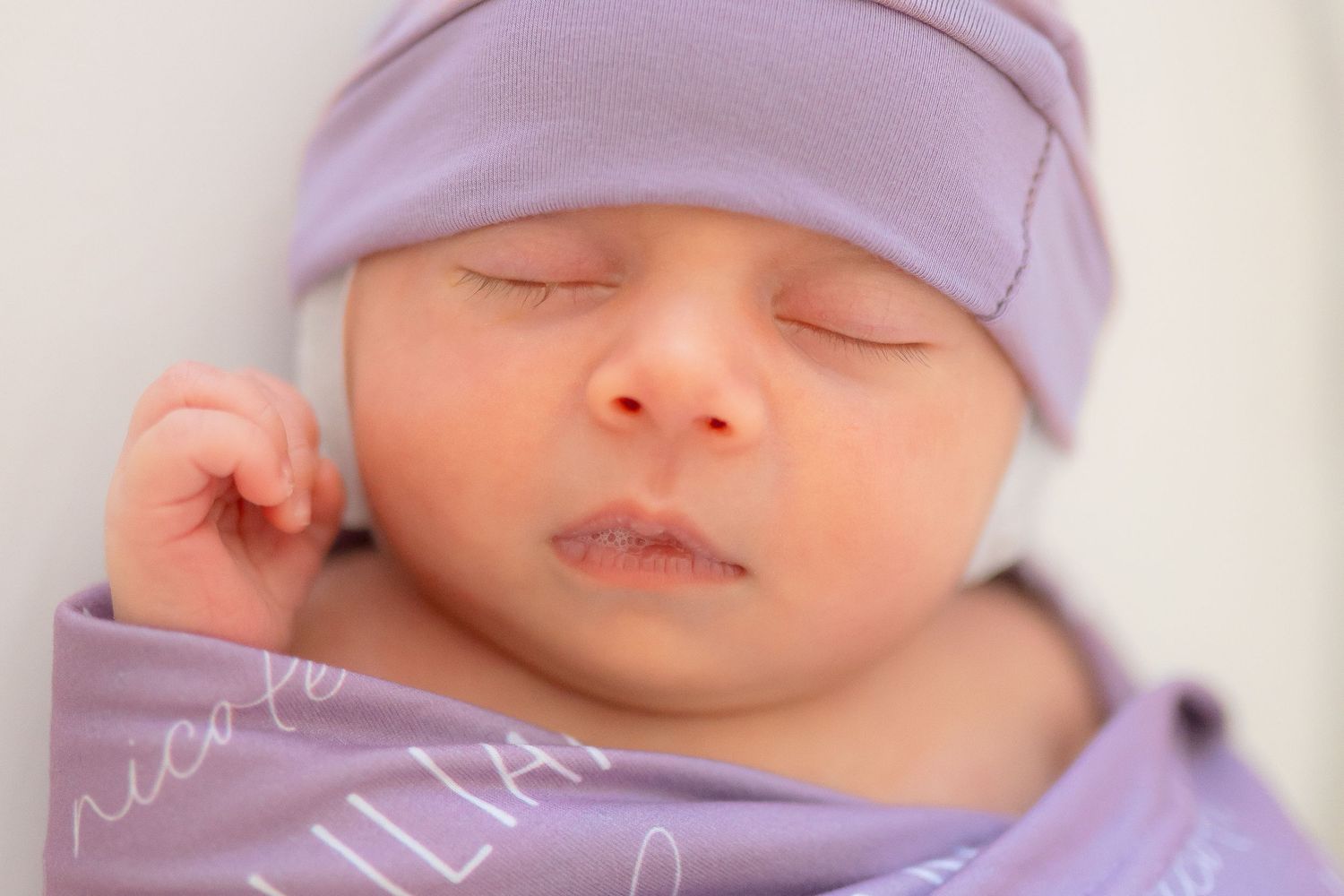 Newborn baby sleeping peacefully wrapped in a personalized purple swaddle blanket with a matching purple knit hat.