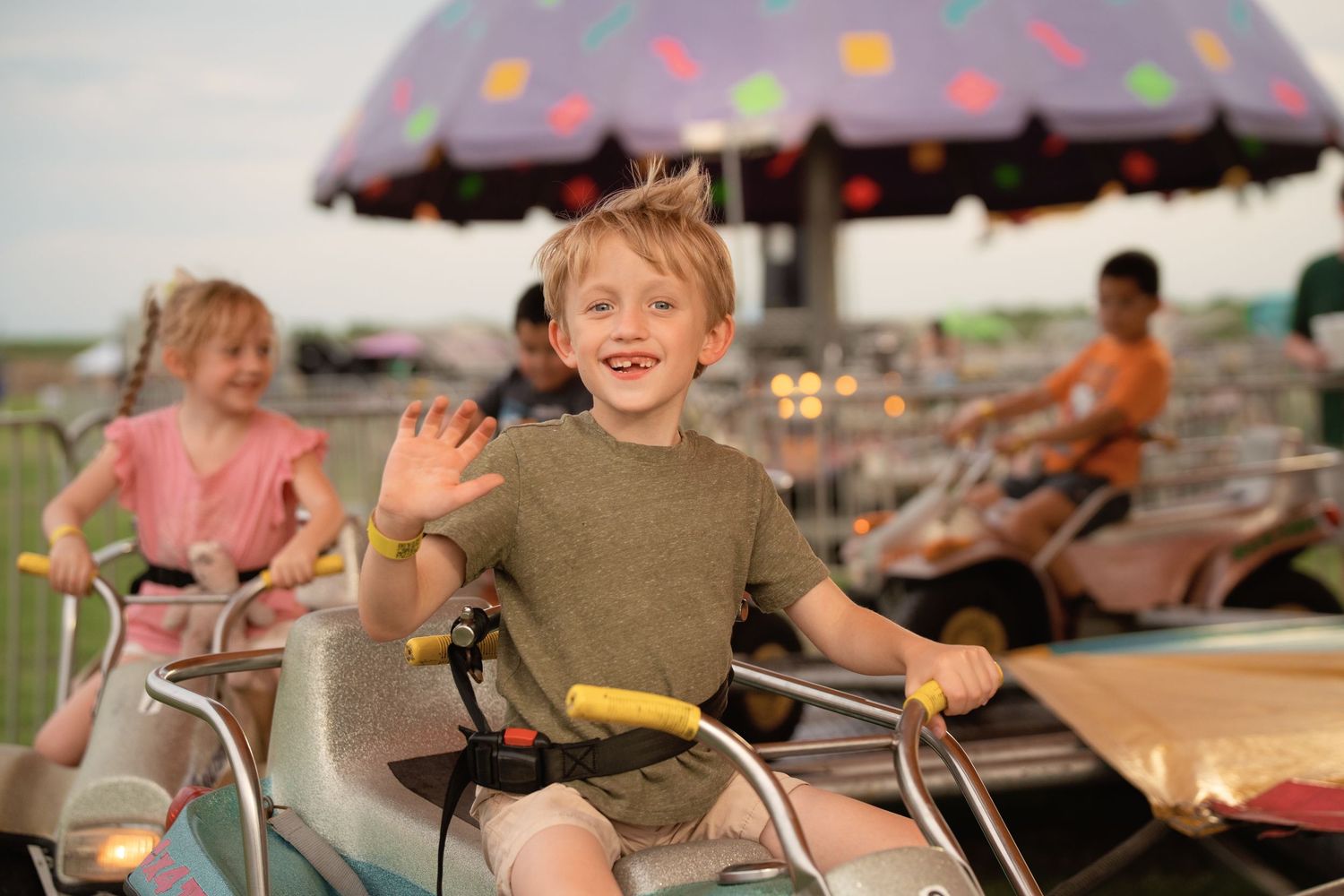 A happy boy waves while riding a carnival ride at an amusement park with other children in the background.