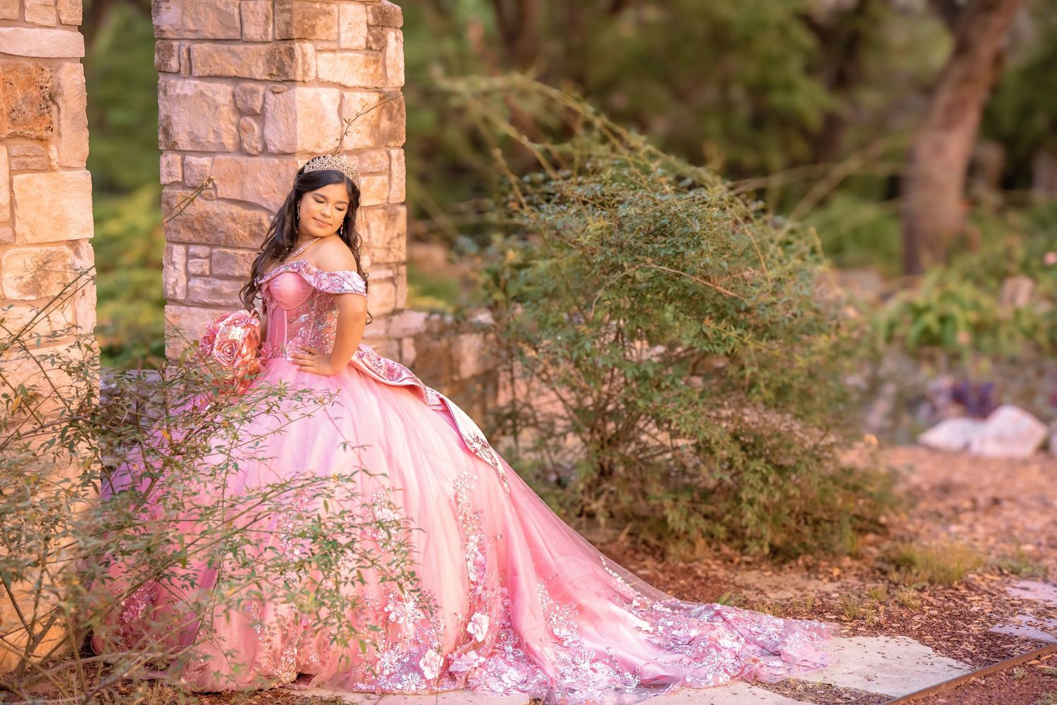 Young woman in a floral pink quinceañera ballgown poses gracefully by a stone pillar in a lush garden setting.