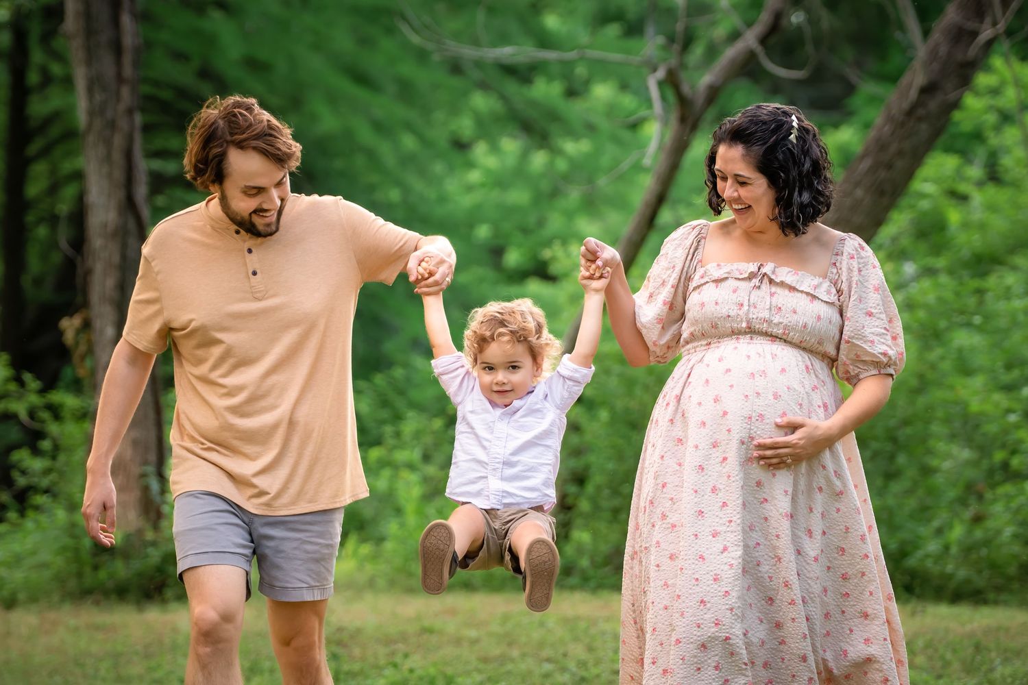 Pregnant woman and man swing toddler boy between them outdoors in green park during maternity photo session.