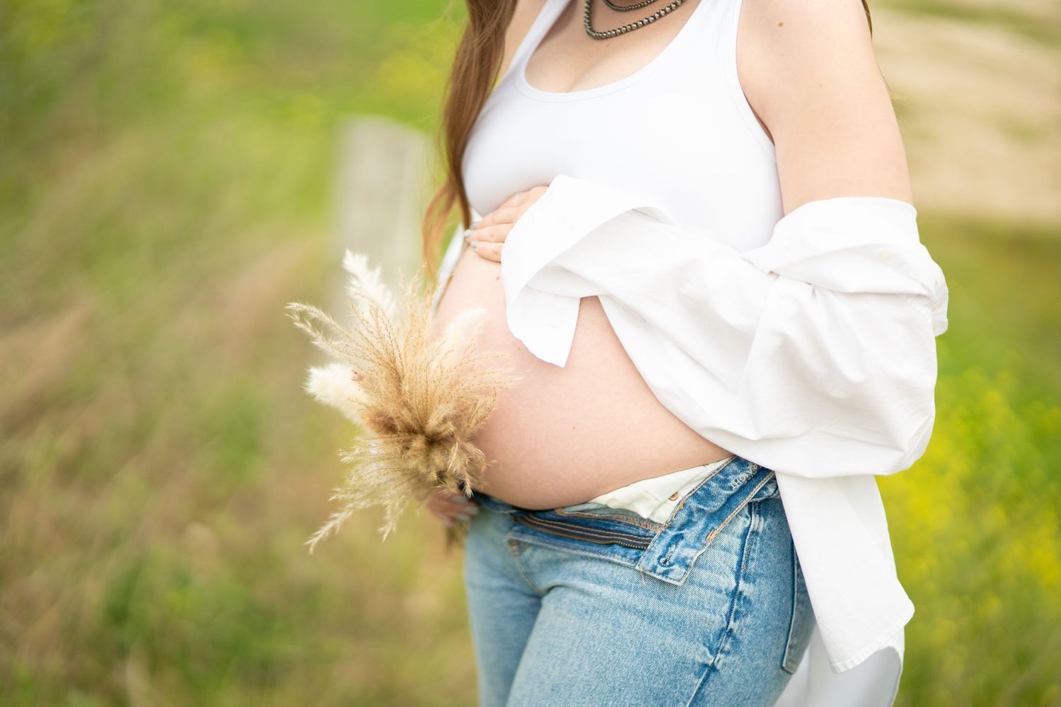 Pregnant woman in white top and jeans holding dried flowers outdoors in a green field.