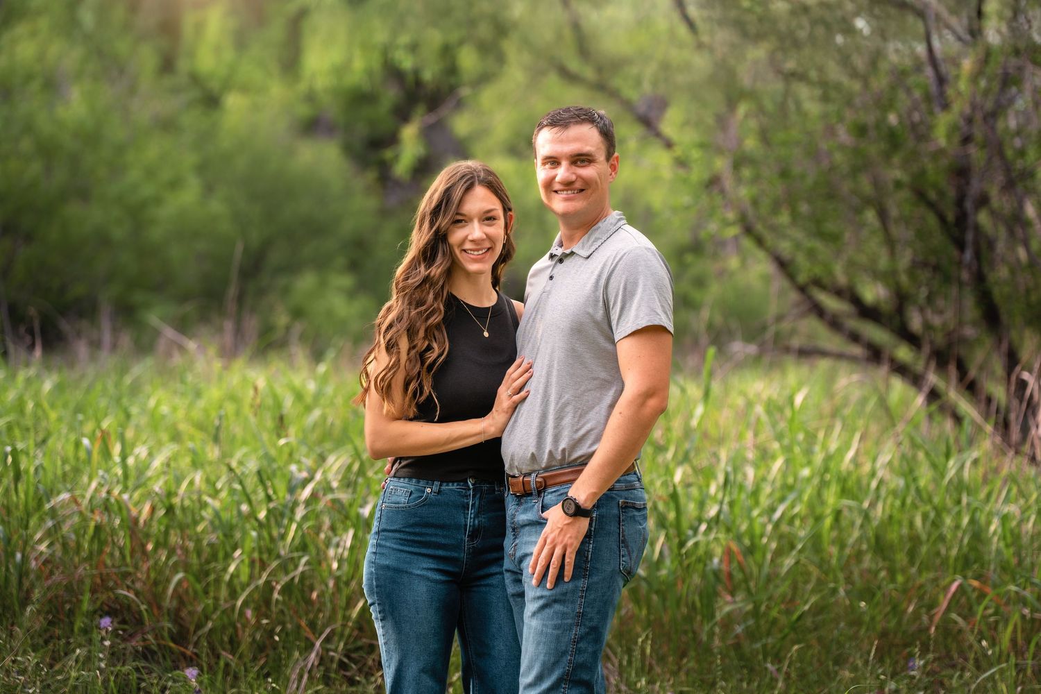 A smiling couple poses together in a lush green outdoor setting, wearing casual jeans and tops.