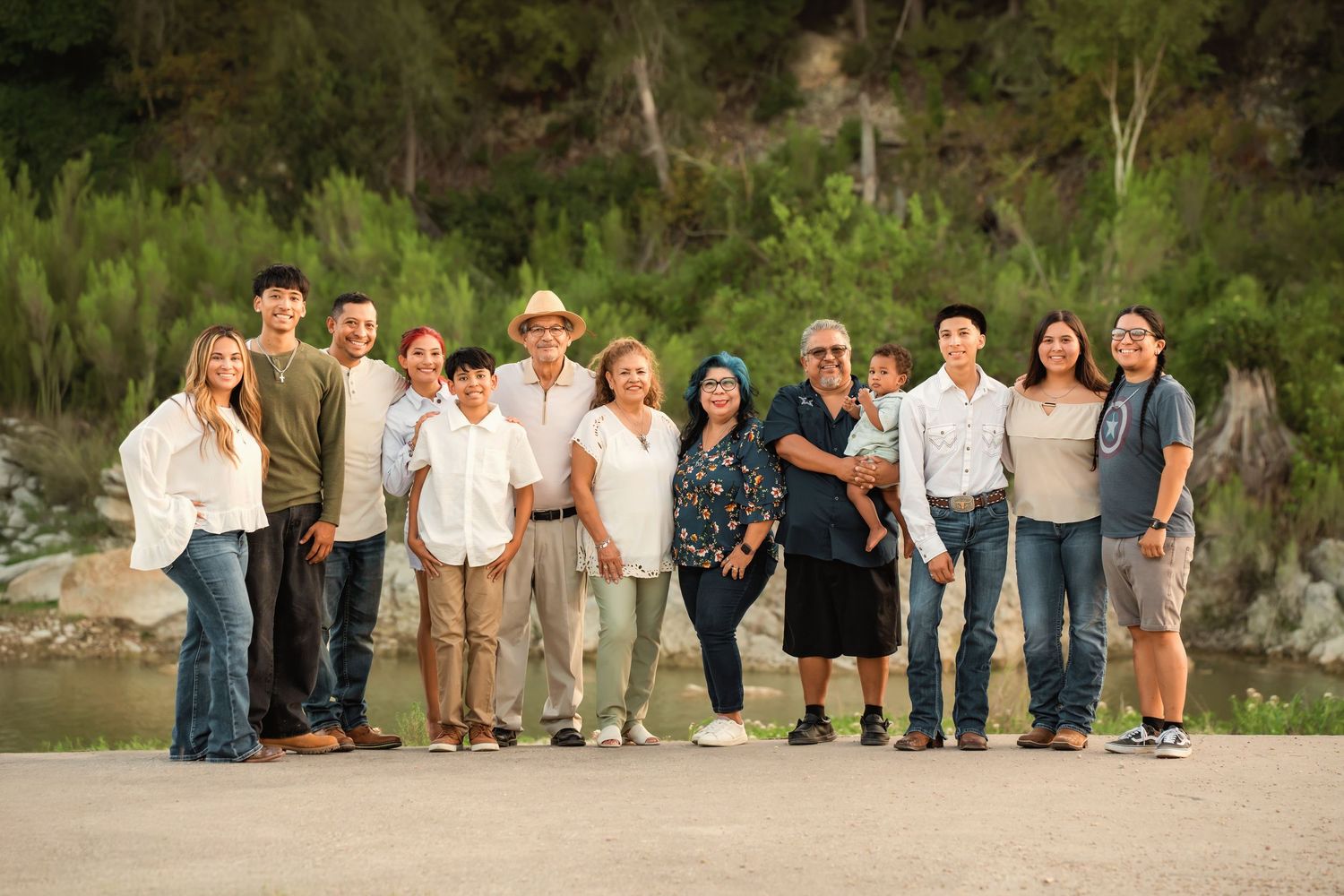 Large multigenerational family group smiling together outdoors near a river with lush green trees in background.