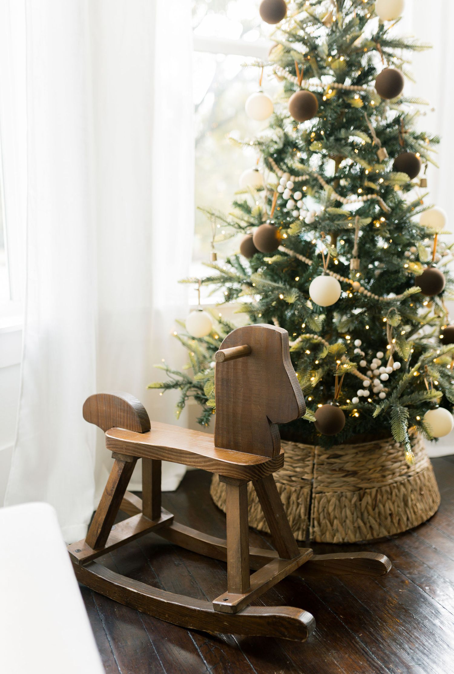Wooden rocking horse toy beside a decorated Christmas tree with gold ornaments and warm lights in a cozy home setting.