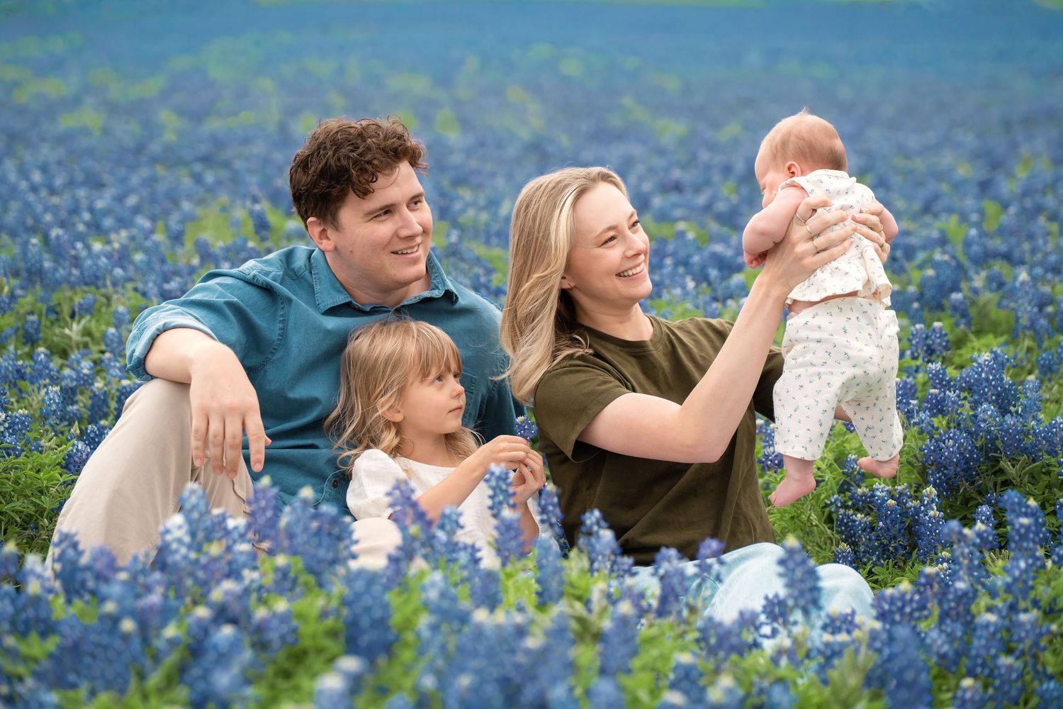 Family of four sitting in a field of blue bluebonnet wildflowers, parents smiling with two young children.