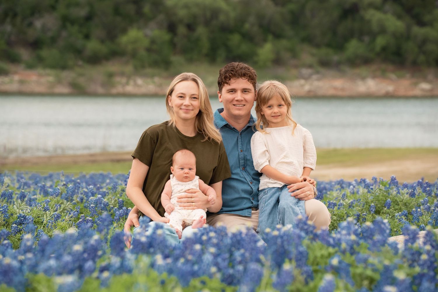 Family of four posing in a field of Texas bluebonnets near a lake, smiling with a baby and young girl.