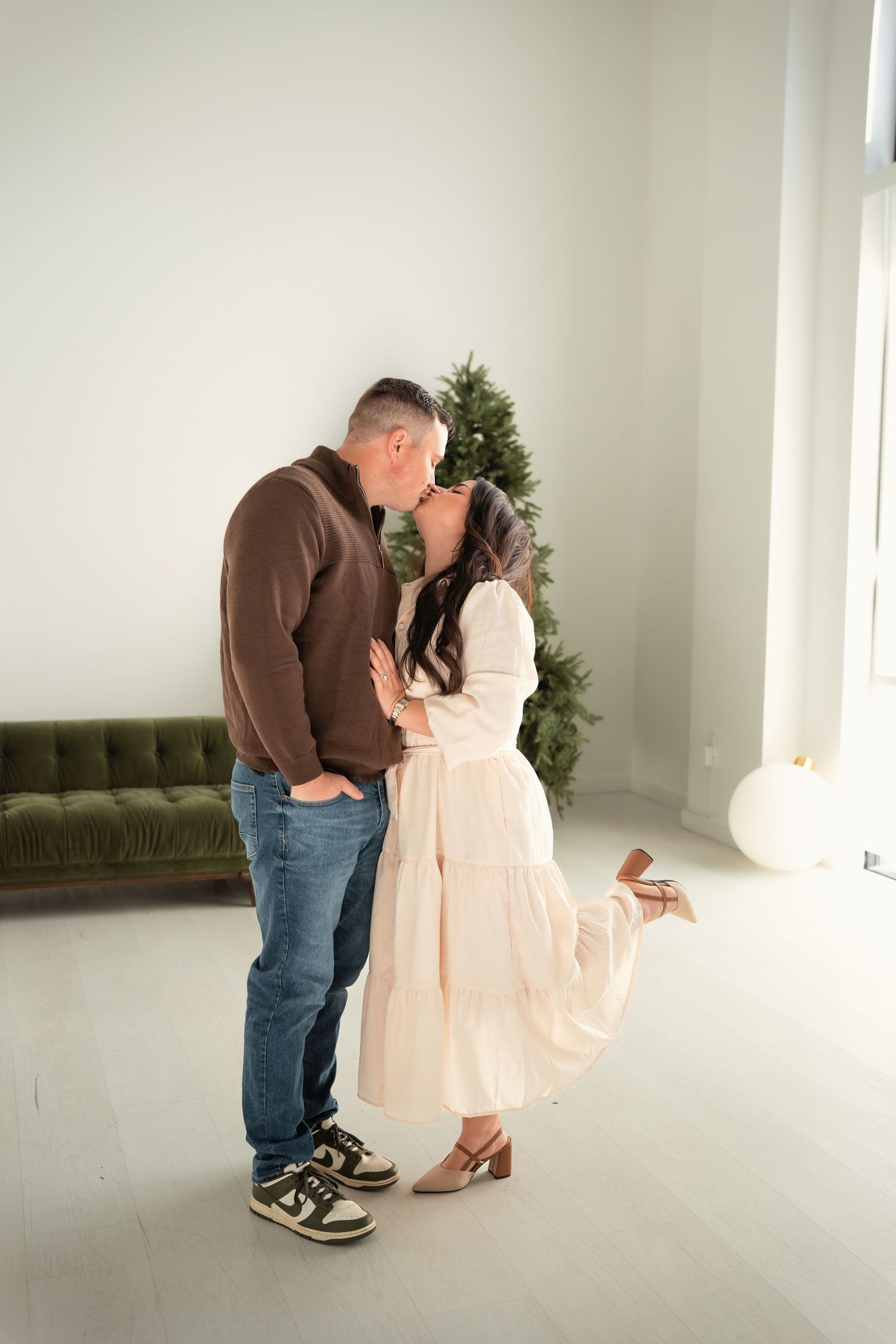 Couple sharing romantic kiss in front of Christmas tree in bright white room with green sofa.