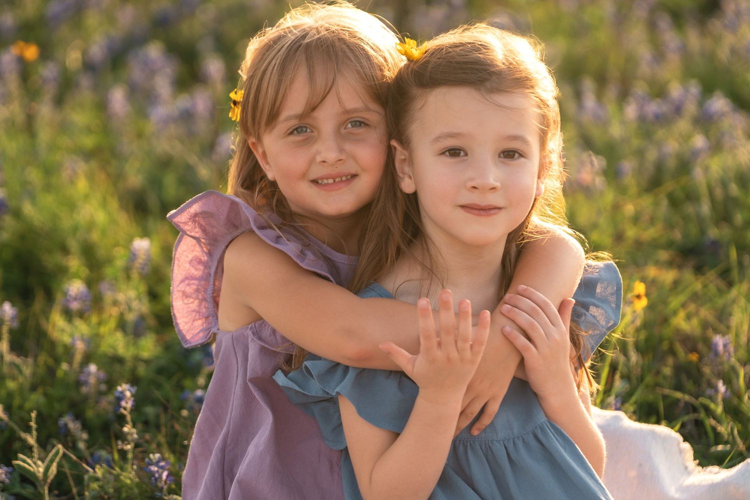 Two young friends in purple and blue dresses embrace while sitting in a sunlit meadow with wildflowers.