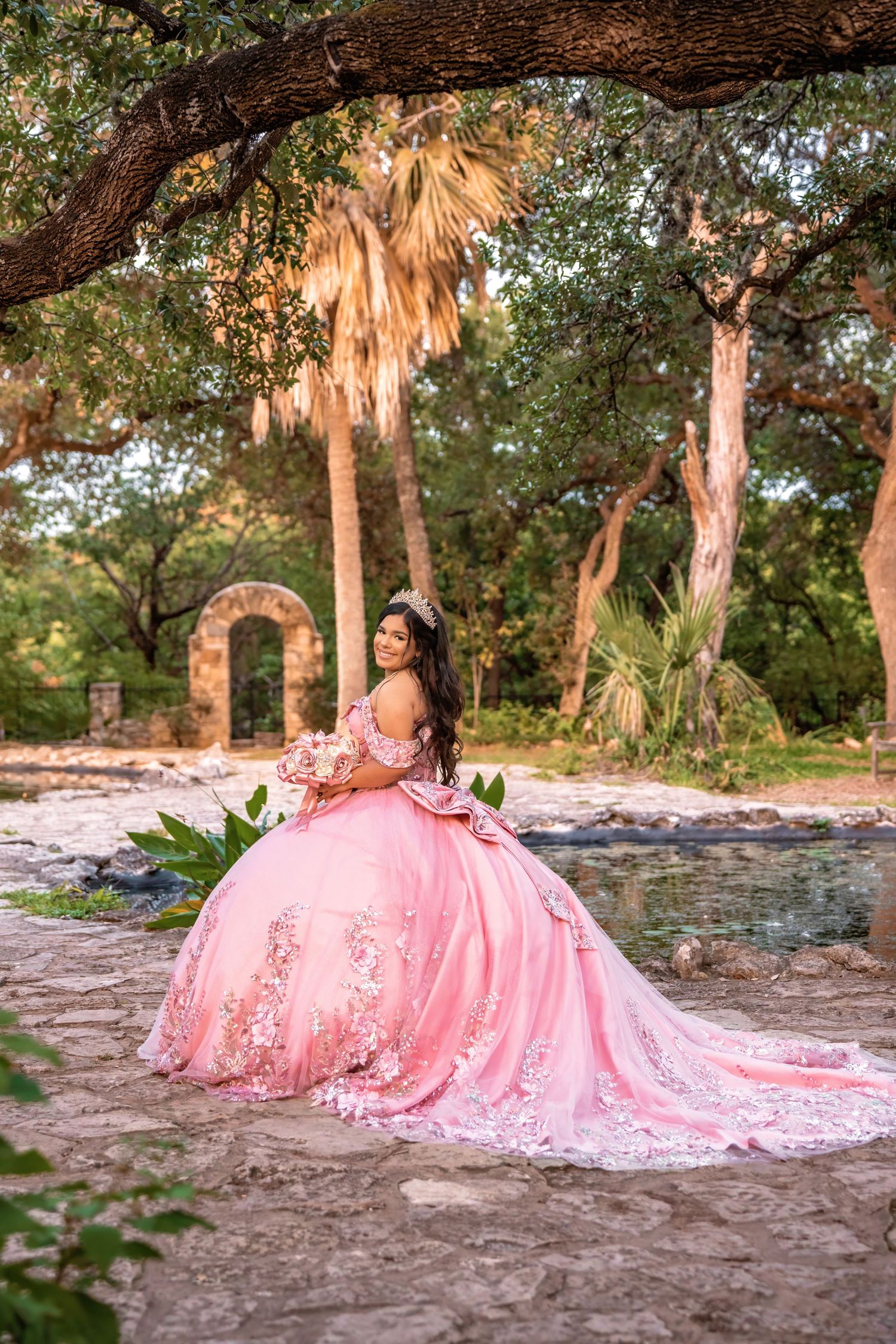 Young woman in a pink ball gown quinceañera dress poses in a lush garden with stone arch and palm trees.