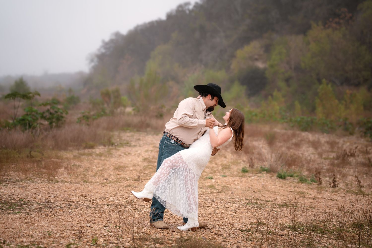 Couple sharing a romantic dip kiss in a rustic outdoor field, man in cowboy hat and woman in white dress.