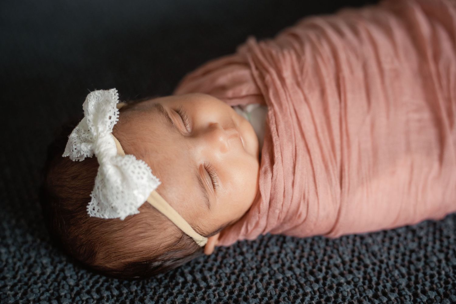 Sleeping newborn baby swaddled in pink wrap with white lace bow headband on gray textured blanket.