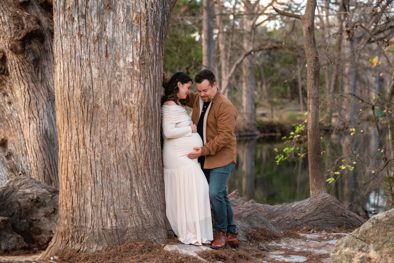 Pregnant couple sharing a tender moment leaning against a large tree outdoors near a peaceful pond.