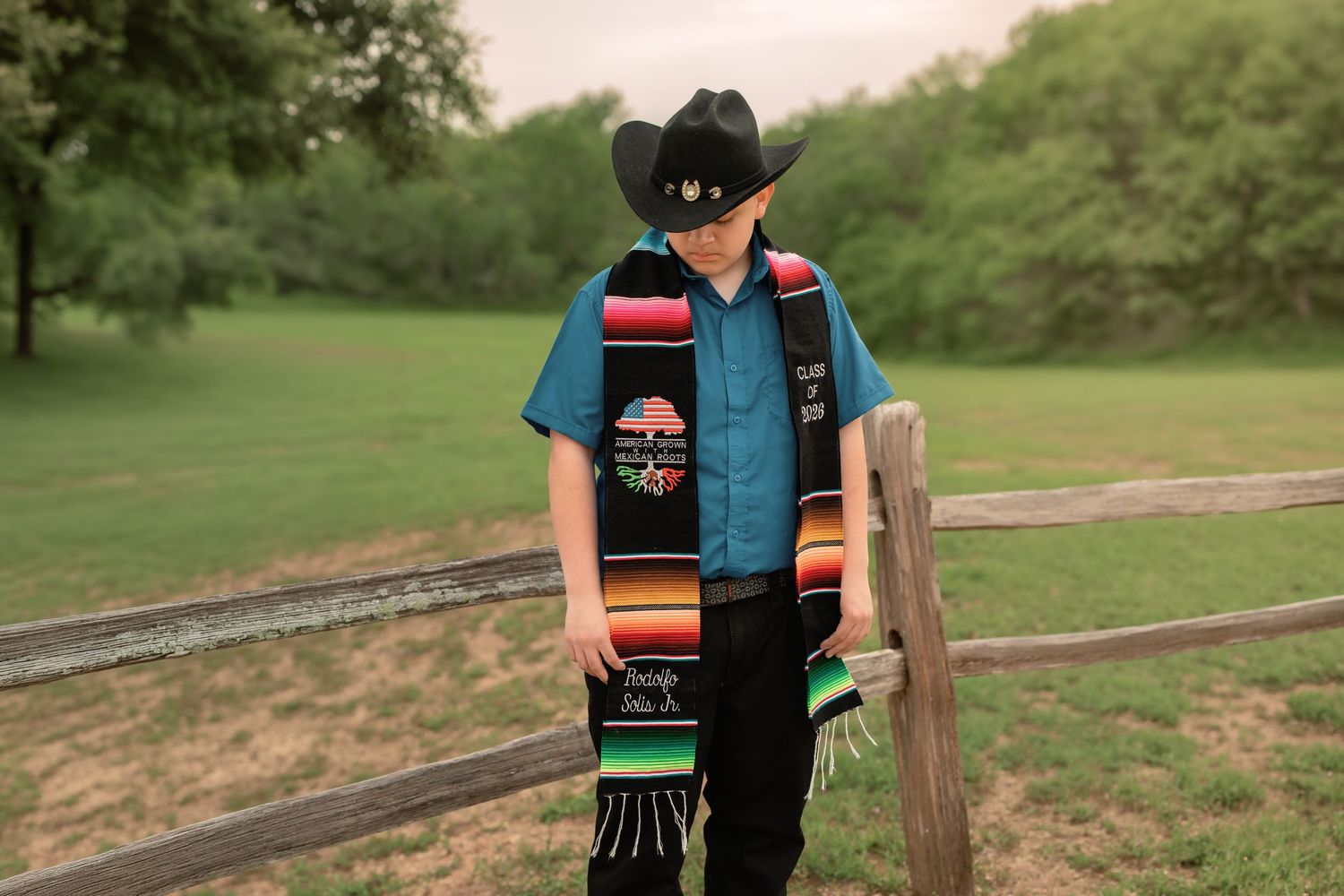 Young boy in black cowboy hat and sunglasses wearing colorful serape graduation stole outdoors.