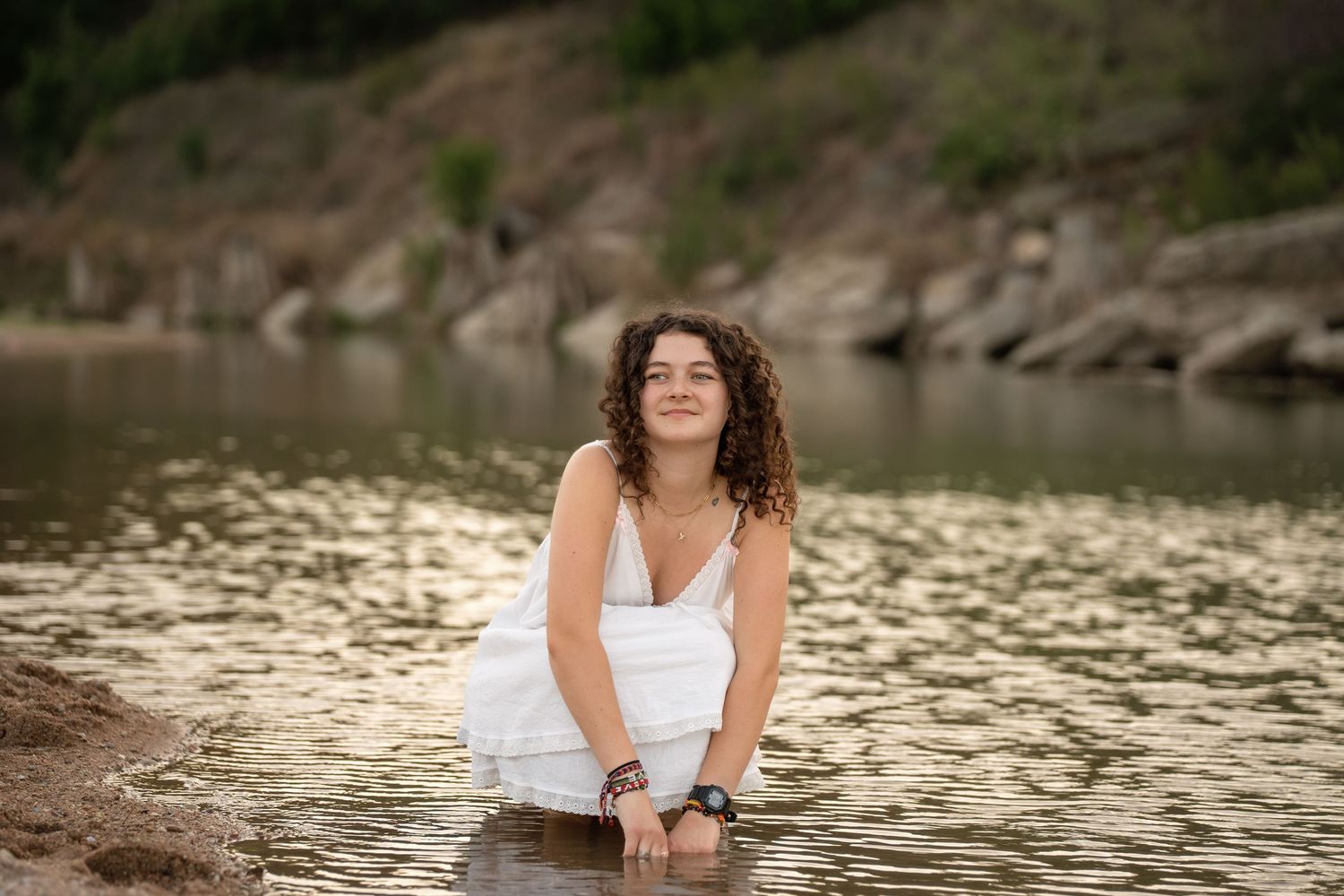 Young woman in white dress sitting in shallow water near rocky shore, enjoying a peaceful outdoor moment.