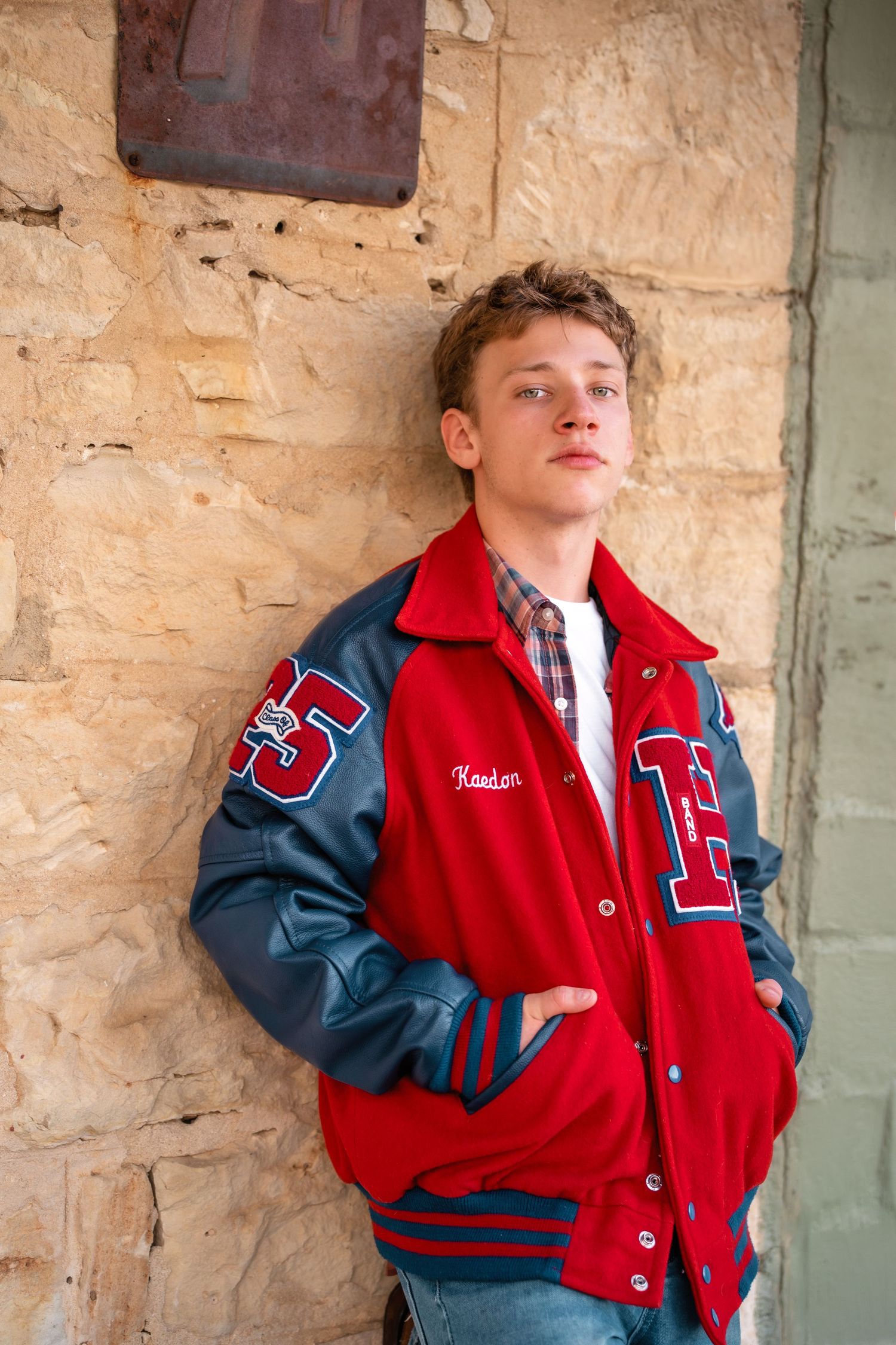 Young man in red letterman jacket leans against stone wall.