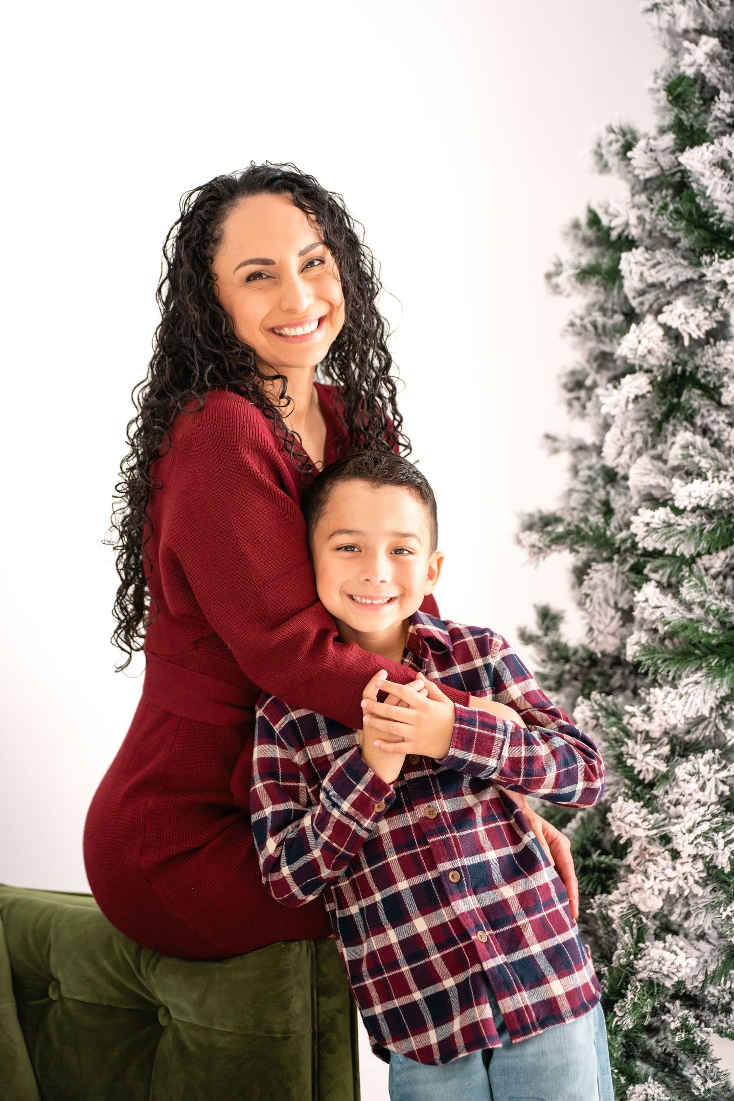 A pair shares a warm embrace and laughter while posing in front of a snowy Christmas tree.