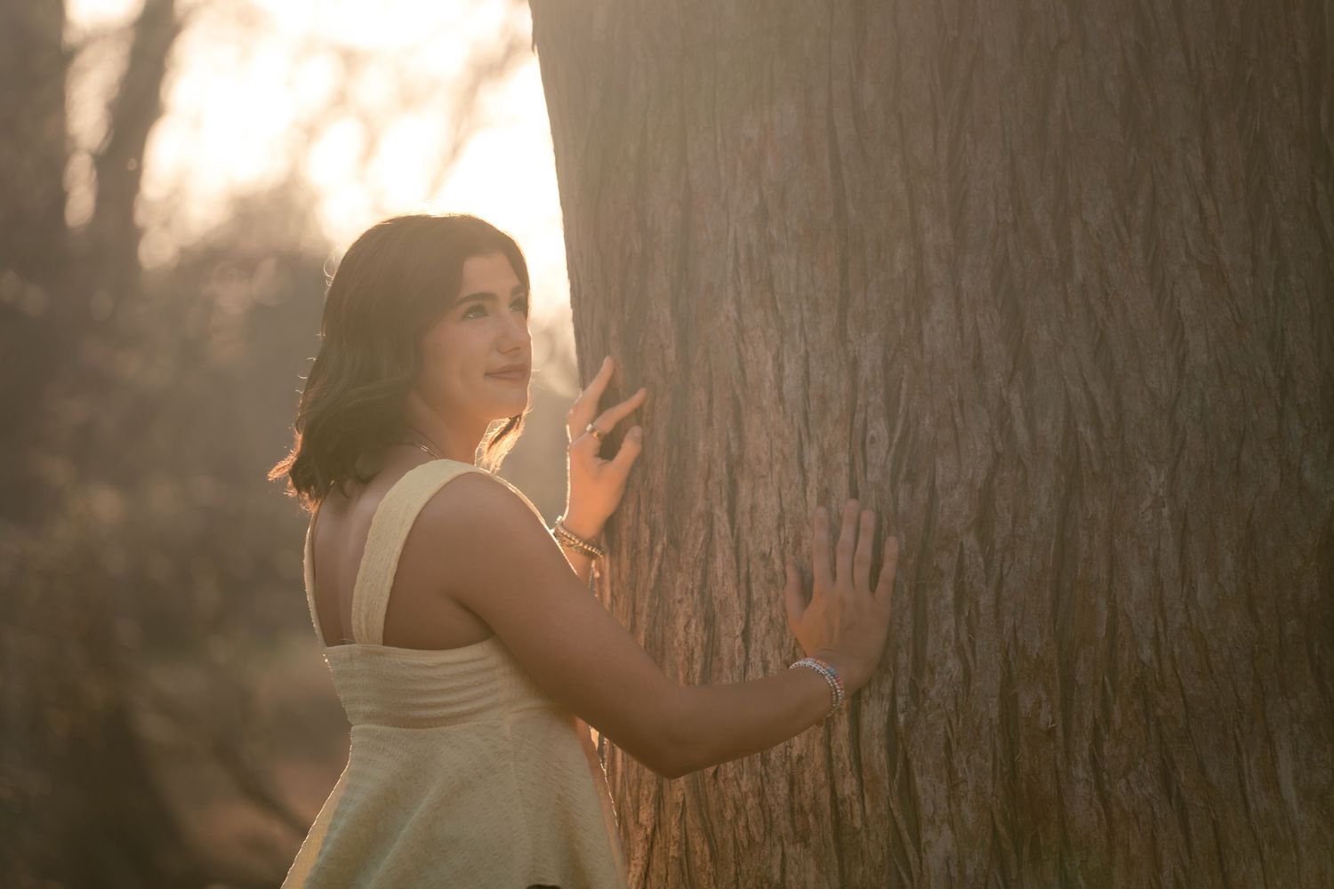 Young woman in white dress leaning against large tree trunk in golden sunset light in a forest.