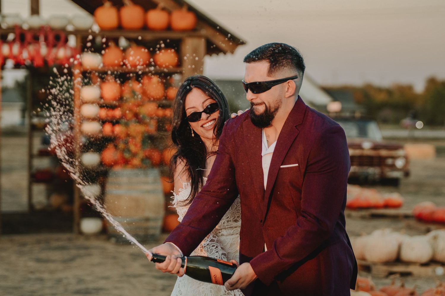 Couple in burgundy blazer and white dress popping champagne at a festive pumpkin patch during golden hour.