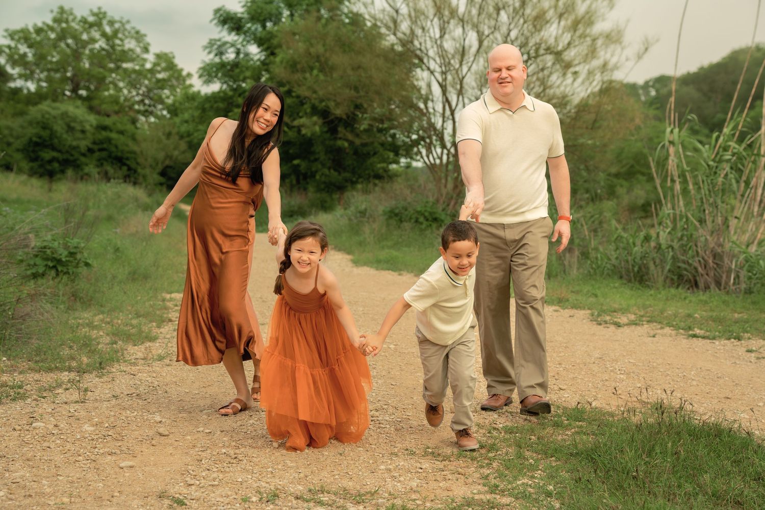 Family of four joyfully walking on a dirt path outdoors, parents holding hands with two young children in autumn-toned outfits.