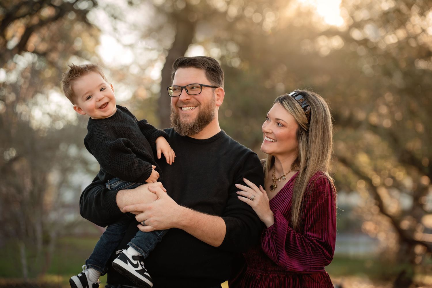 A happy family of three posing outdoors, father holding toddler while mother smiles beside them in warm golden light.