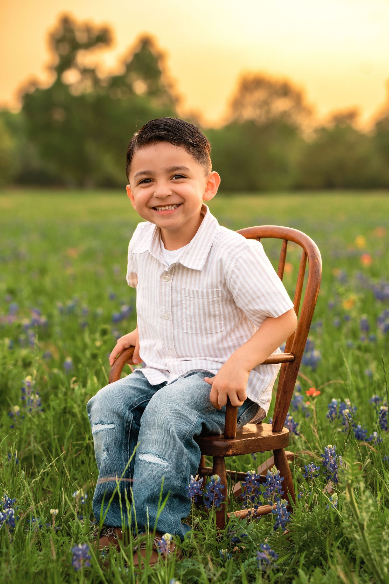 A child sits in a wooden chair among blooming bluebonnets during golden hour sunset in a Texas field.