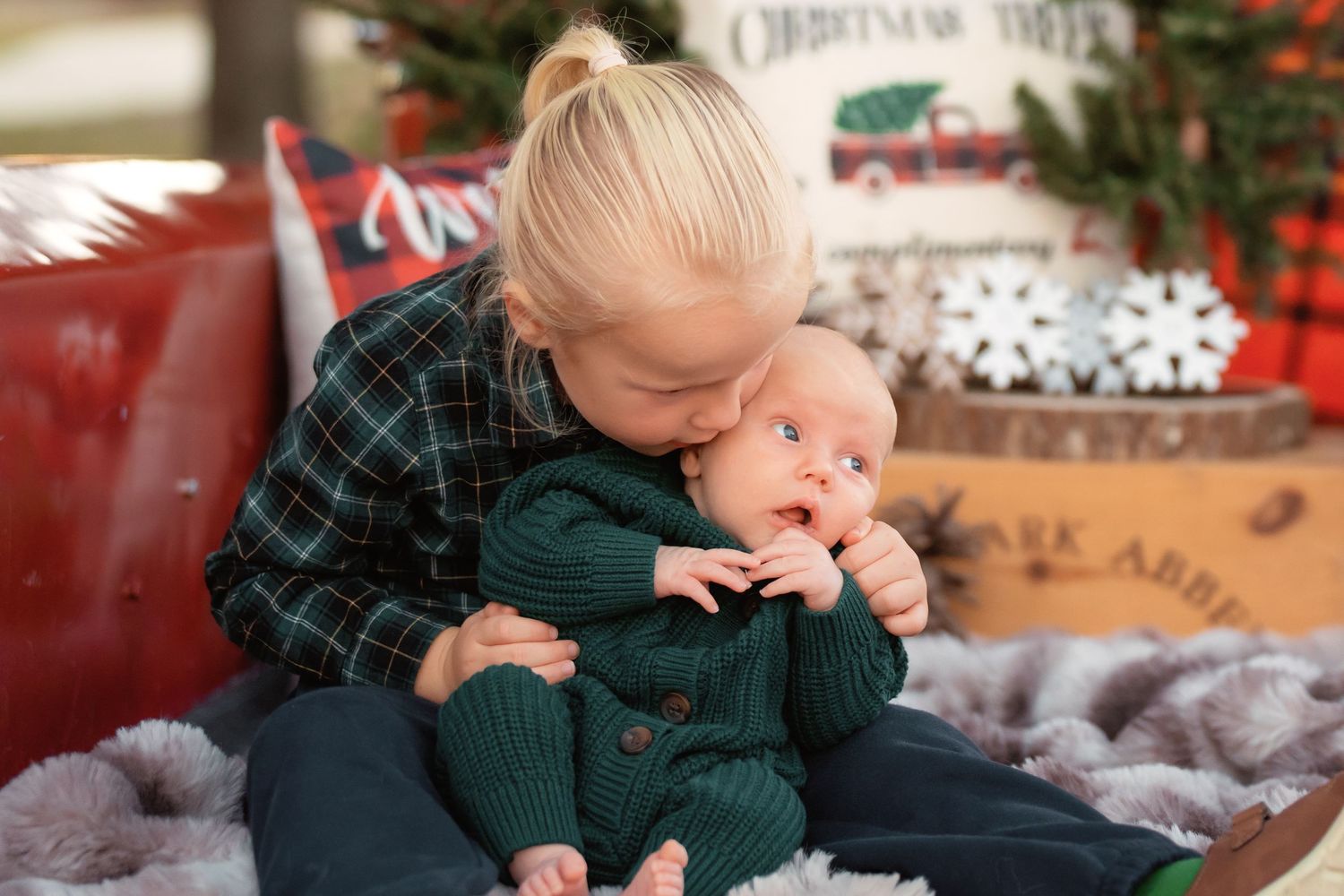Older toddler girl kissing newborn baby in green outfits during a Christmas holiday photo session.