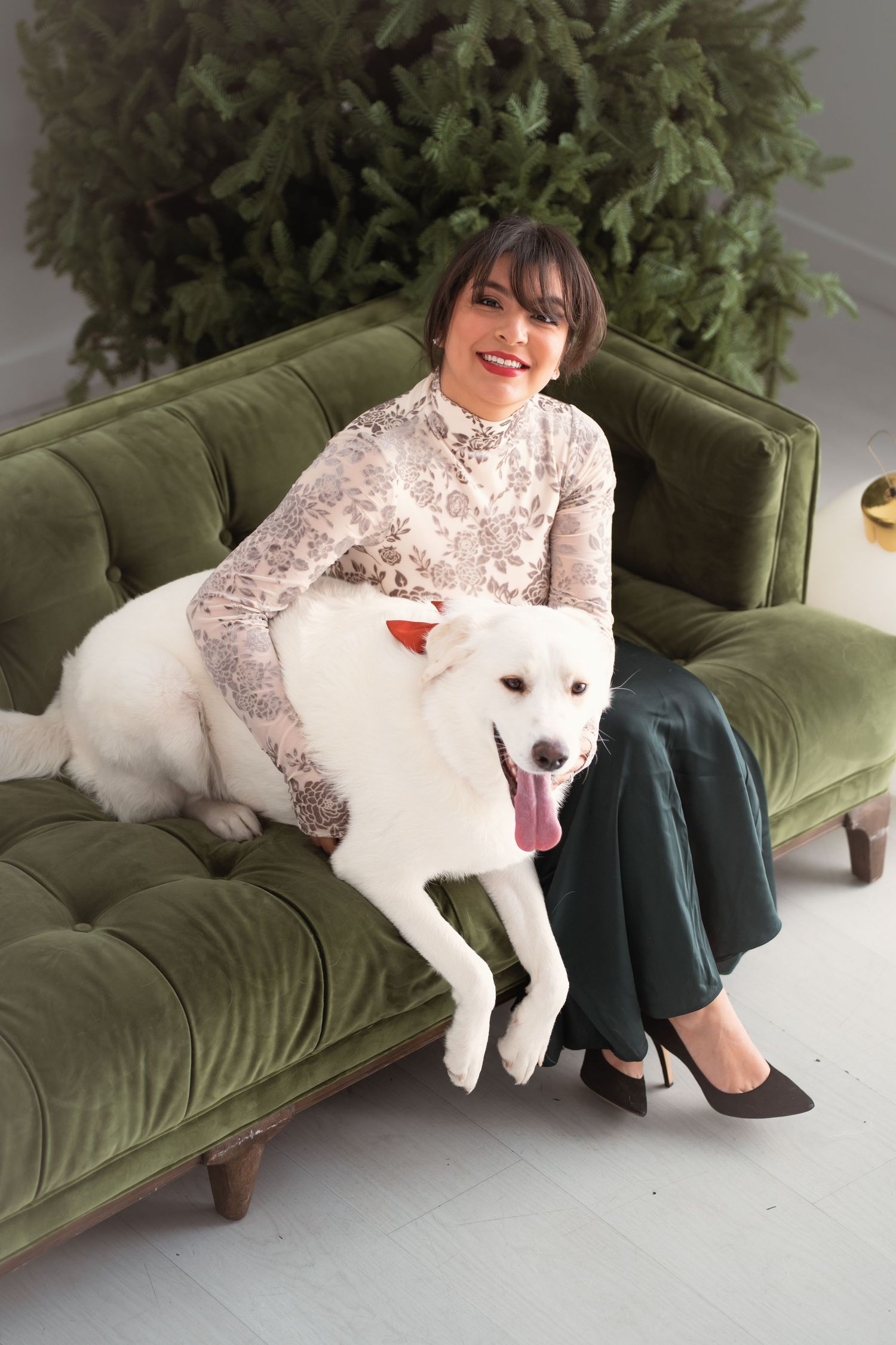 Smiling woman sitting on green velvet sofa with white dog in front of Christmas tree.