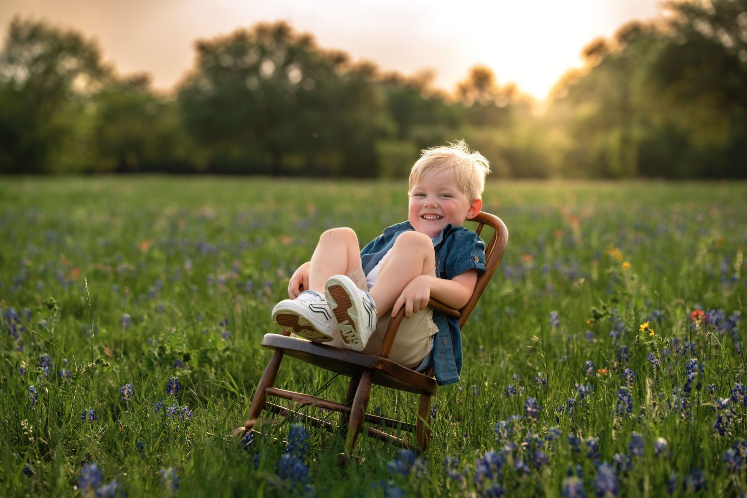 A person sits in a wooden chair surrounded by wildflowers and tall grass during golden hour at sunset.