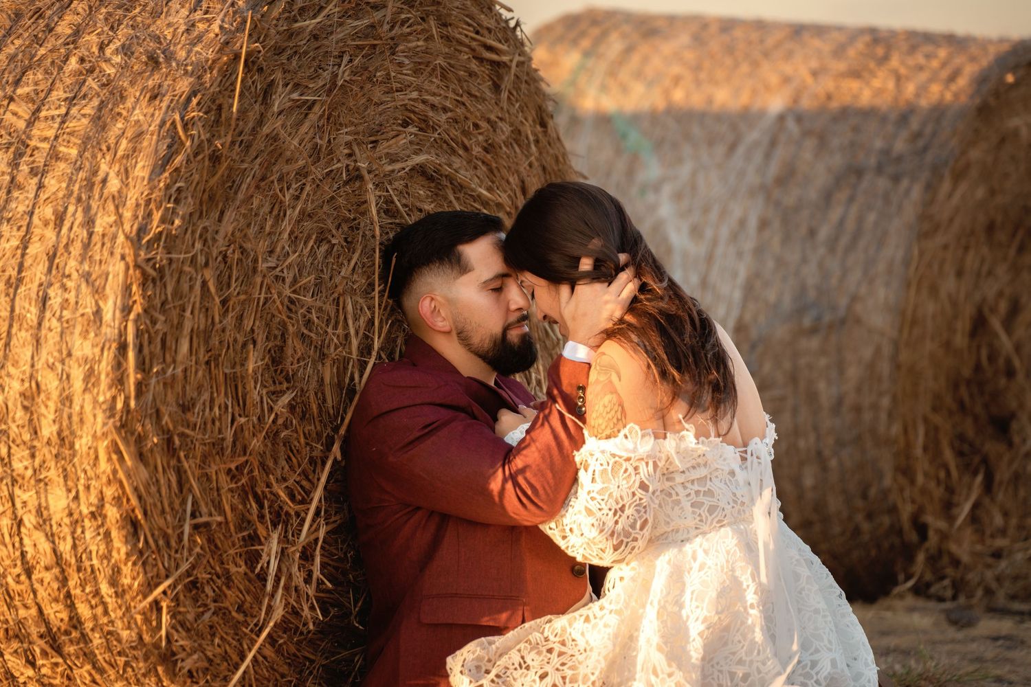 A couple shares an intimate moment in front of golden hay bales during sunset.