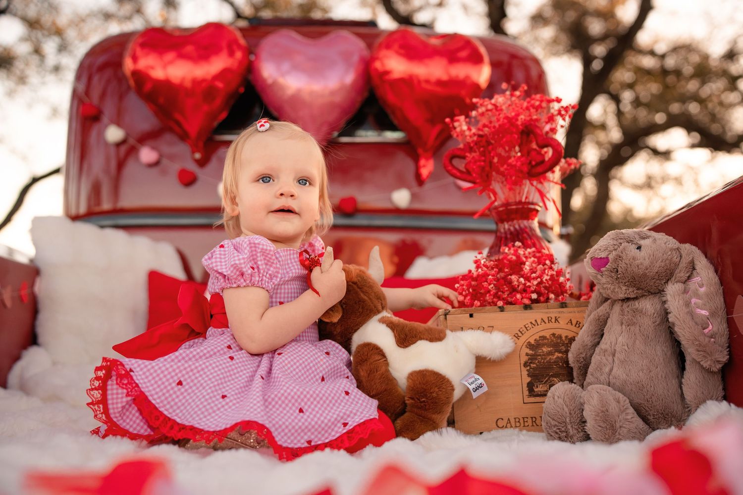 Baby girl in a pink polka dot dress sits on a white blanket with plush bunnies, red heart balloons and Valentine decorations.