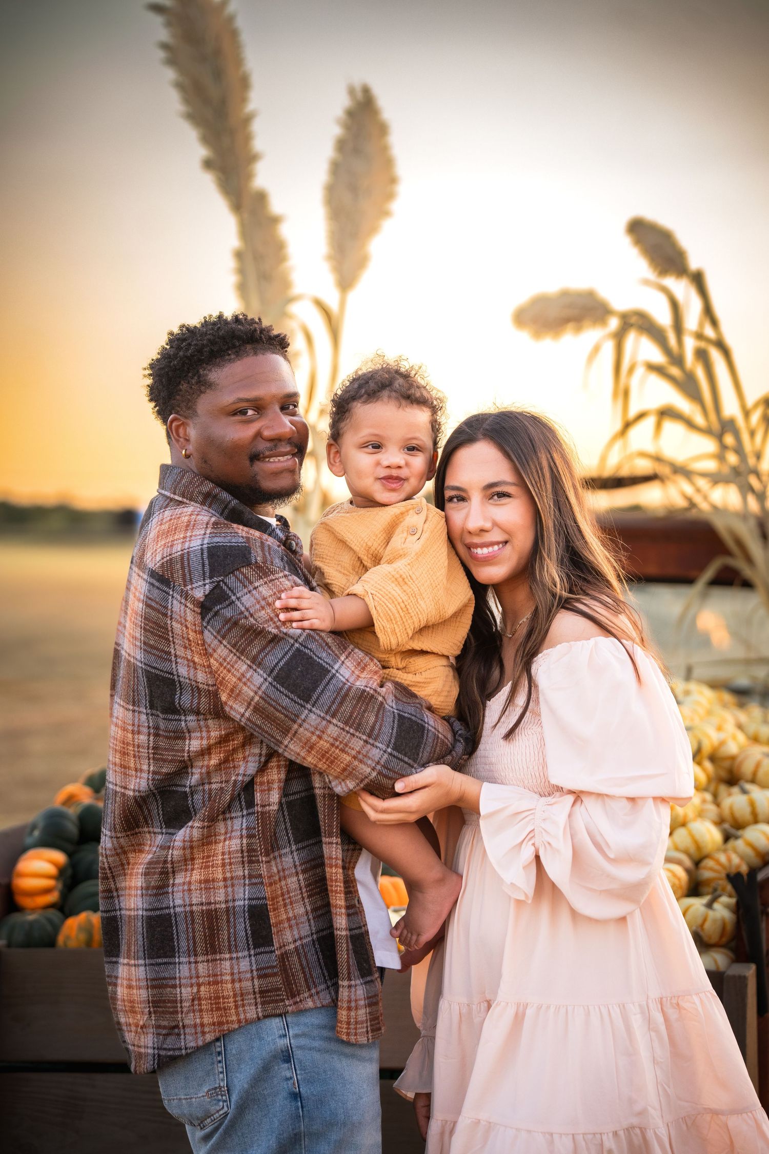 A family embraces in a pumpkin patch at sunset while wearing casual fall clothing and enjoying the golden hour.