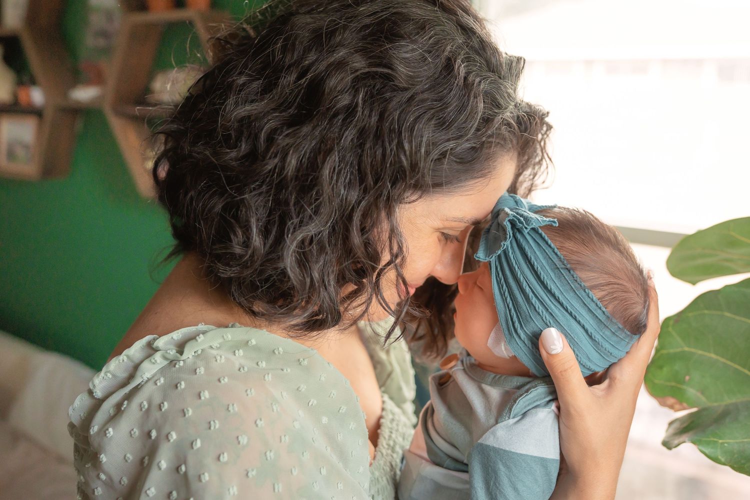 A curly-haired mother tenderly touches foreheads with her newborn baby wrapped in a blue swaddle.