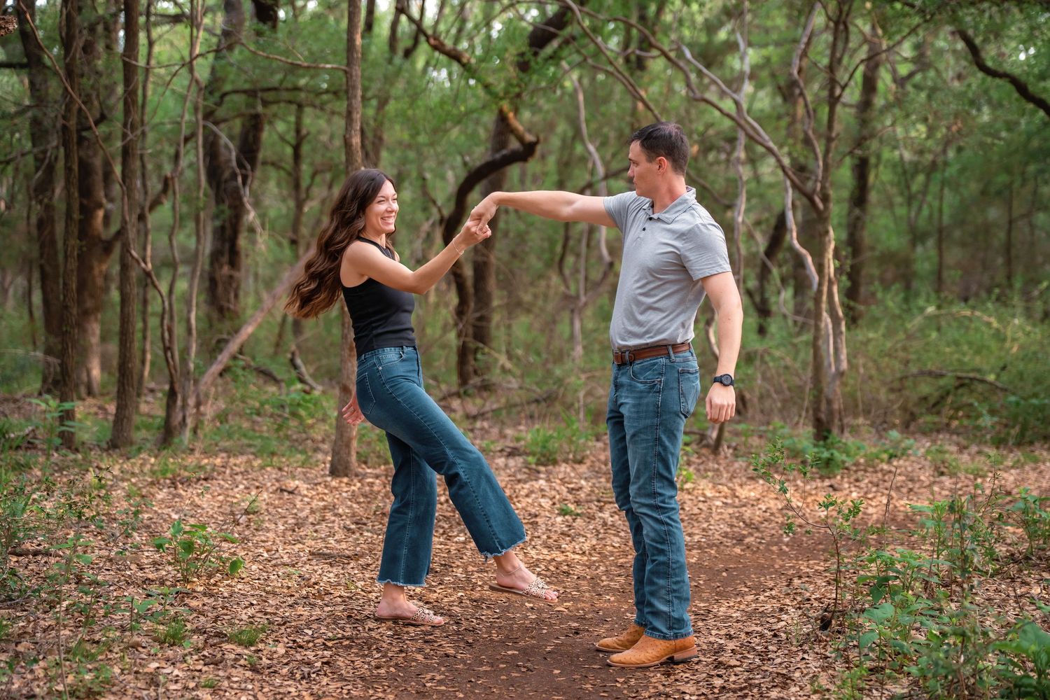 Couple dancing joyfully in a lush forest, holding hands and spinning on a dirt path surrounded by trees.