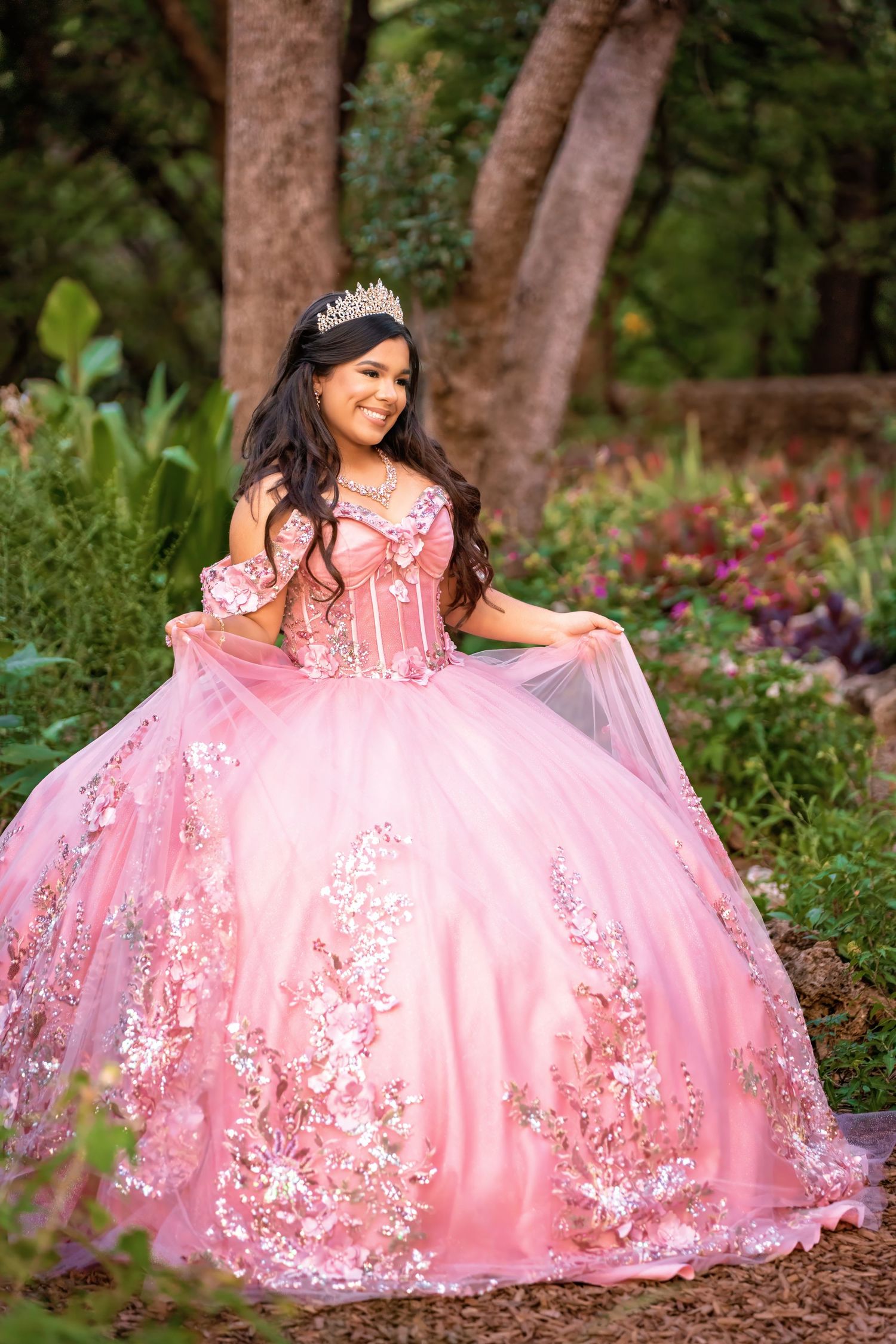 Young woman in pink floral quinceañera ballgown with tiara poses in lush garden setting.