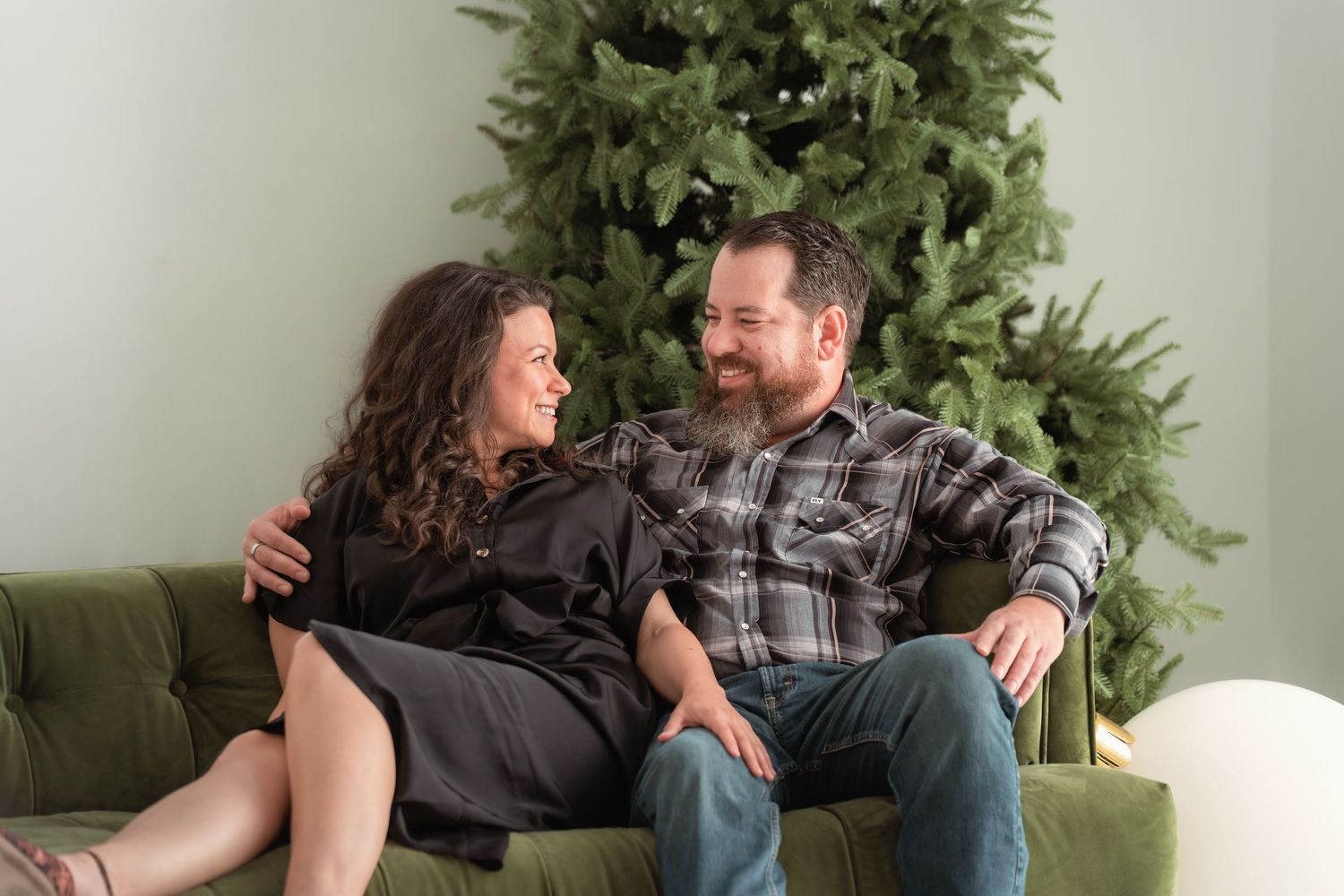 Couple smiling at each other on green velvet sofa in front of decorated Christmas tree holiday portrait.