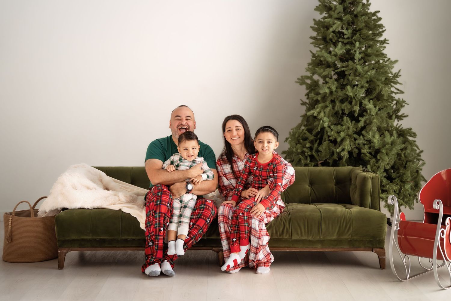 Family of four in matching Christmas pajamas sitting on green velvet sofa beside decorated holiday tree.