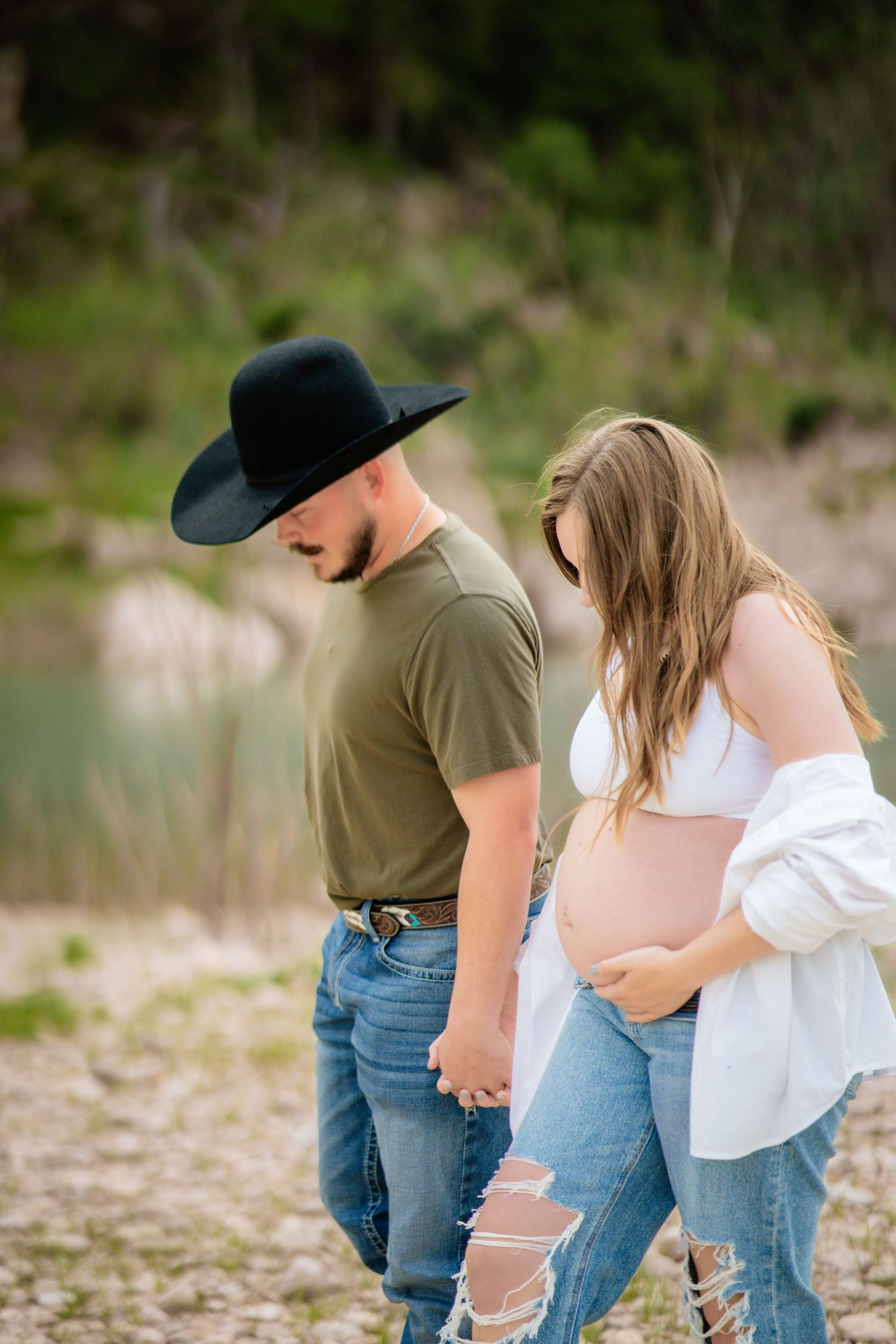 Couple holding hands outdoors, man in cowboy hat and green shirt, pregnant woman in white top and ripped jeans.