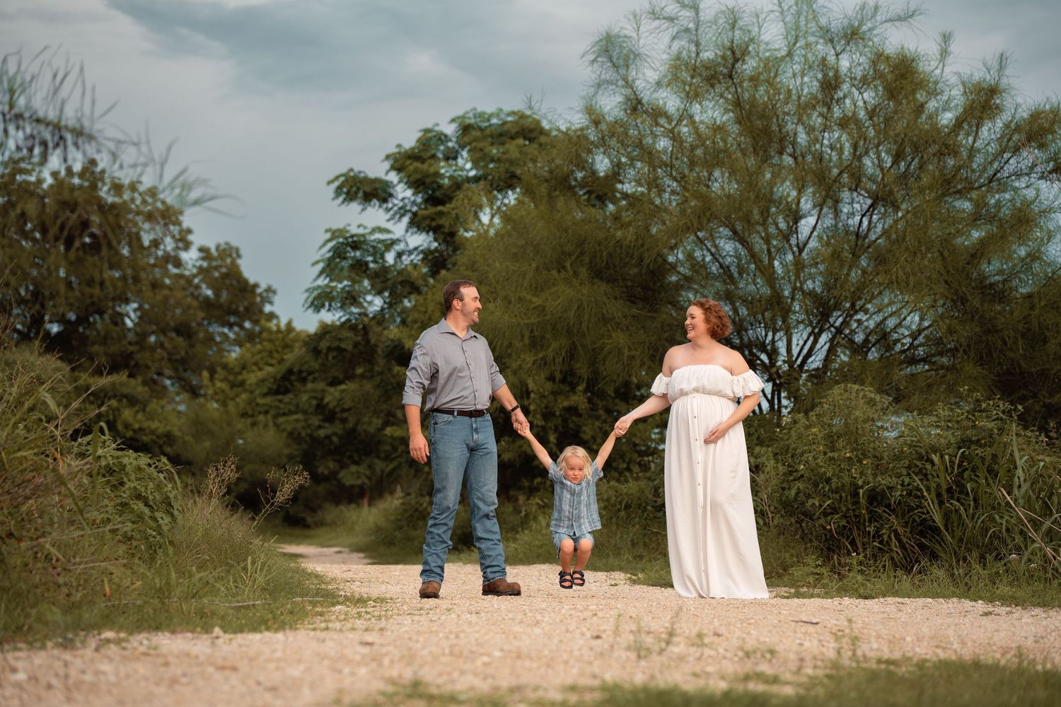 Pregnant woman in white dress holds hands with toddler and partner on a scenic outdoor path surrounded by lush greenery.