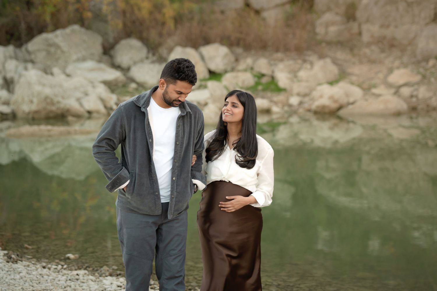 Pregnant couple standing by a serene lake with rocky shores, woman cradling baby bump in white sweater and brown skirt.