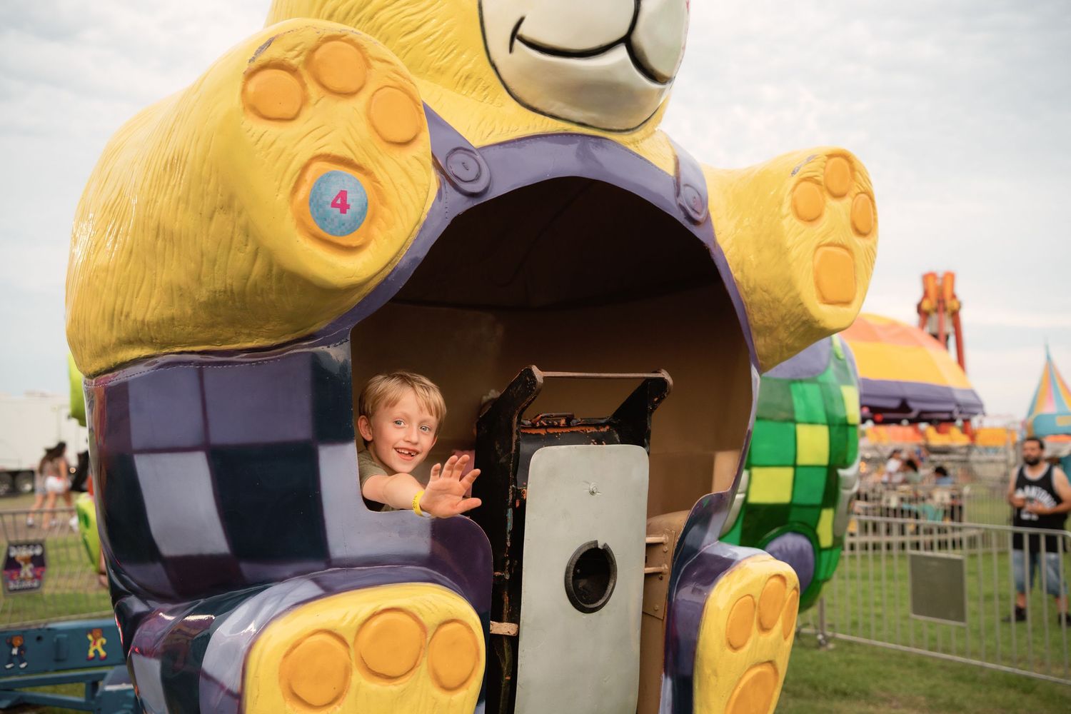 Young child smiling inside a colorful bear-shaped carnival ride at an outdoor fairground.